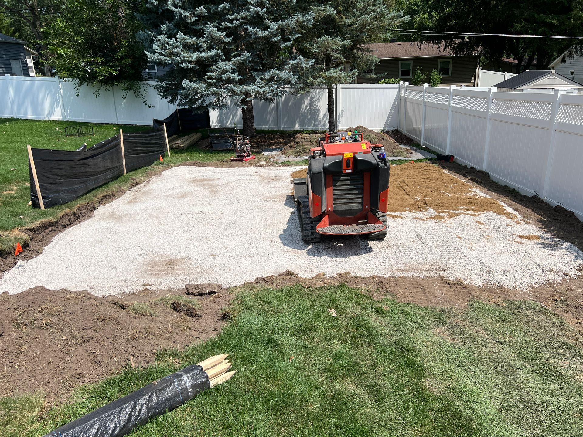 An orange skid-steer loader sits on a prepared gravel base in a residential backyard, surrounded by a white vinyl fence.