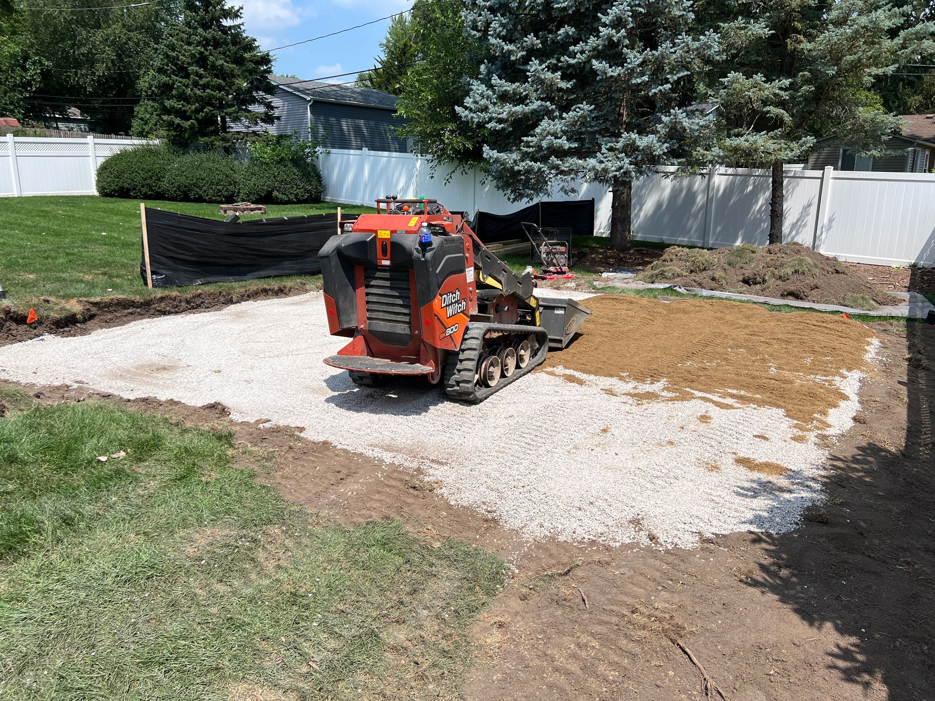 An orange compact track loader sits on a gravel patch in a backyard during a landscaping project.