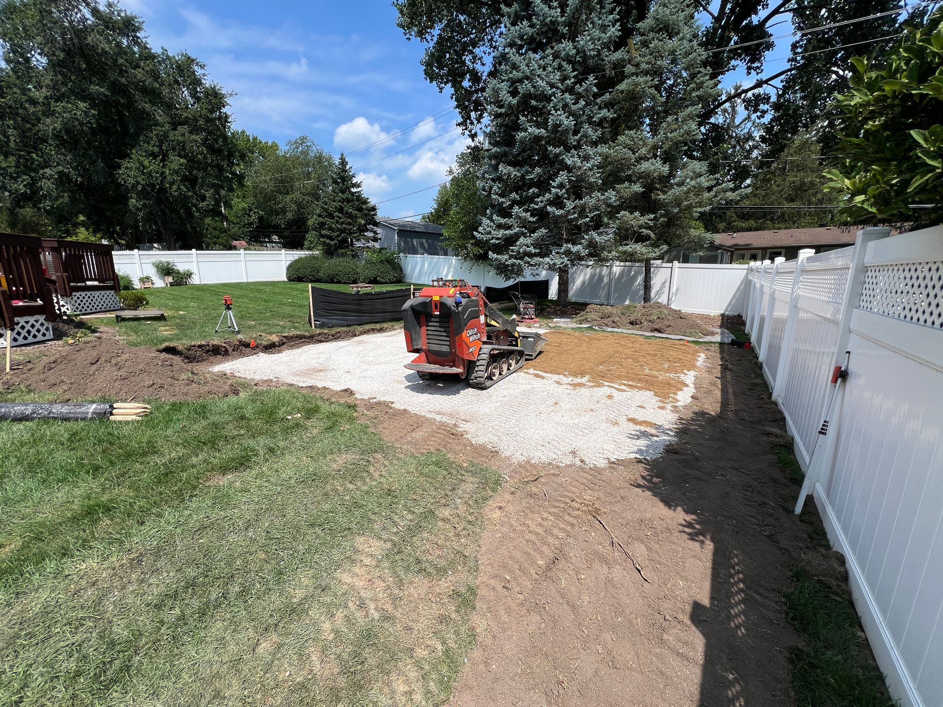 A small construction vehicle sits on a gravel base in a residential backyard undergoing landscaping work.