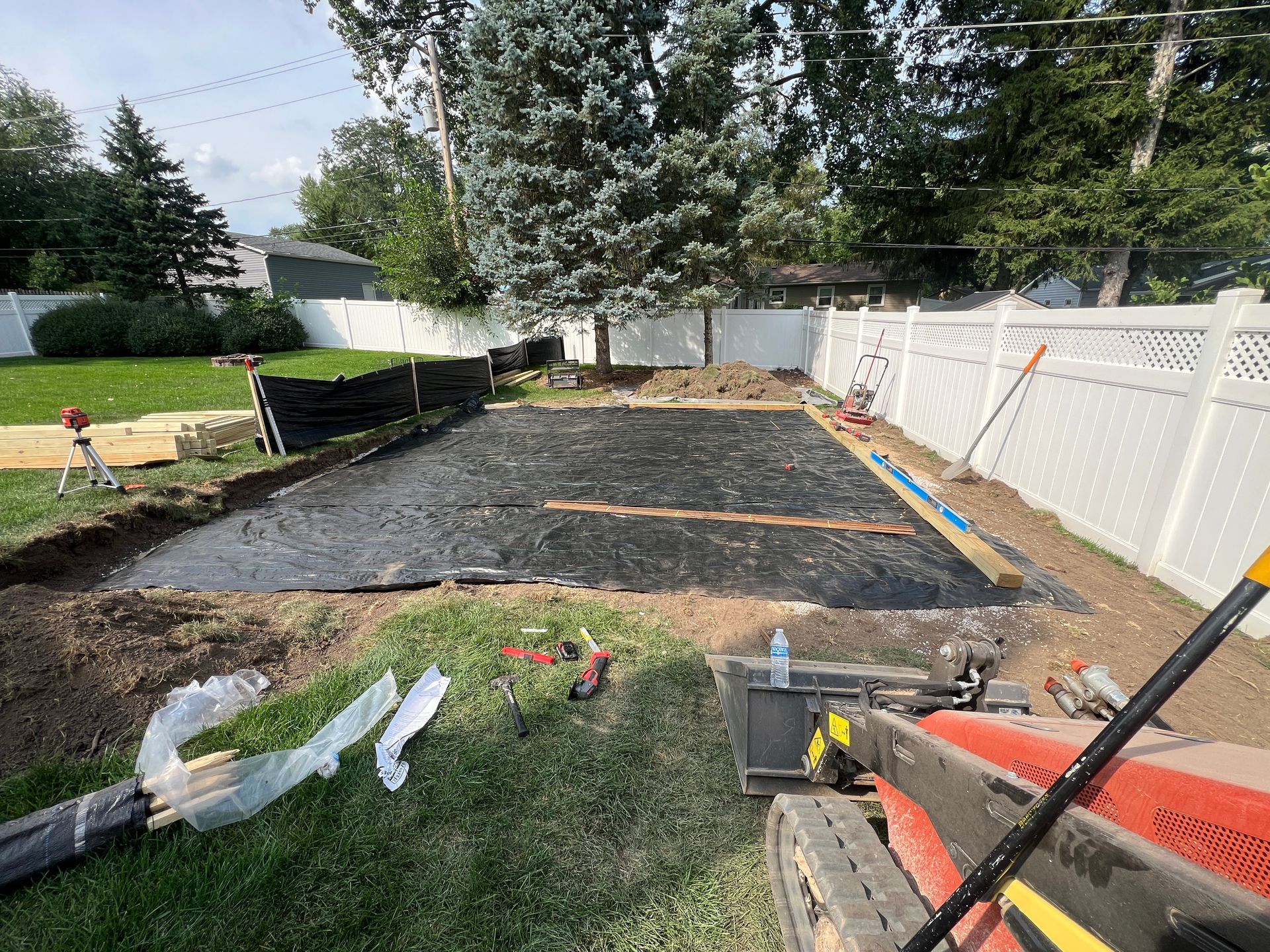 A construction site in a backyard shows a rectangular area covered with black landscape fabric, framed by wooden boards.