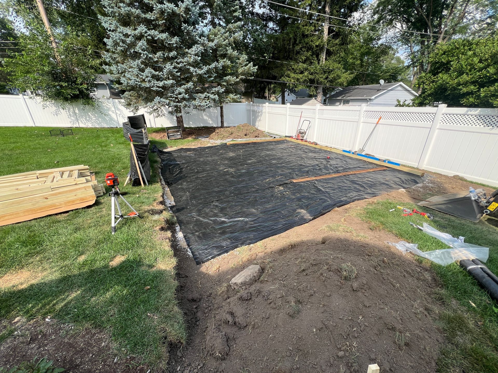 A rectangular patch of ground covered in black landscape fabric in a backyard, beside a white vinyl fence and lawn.