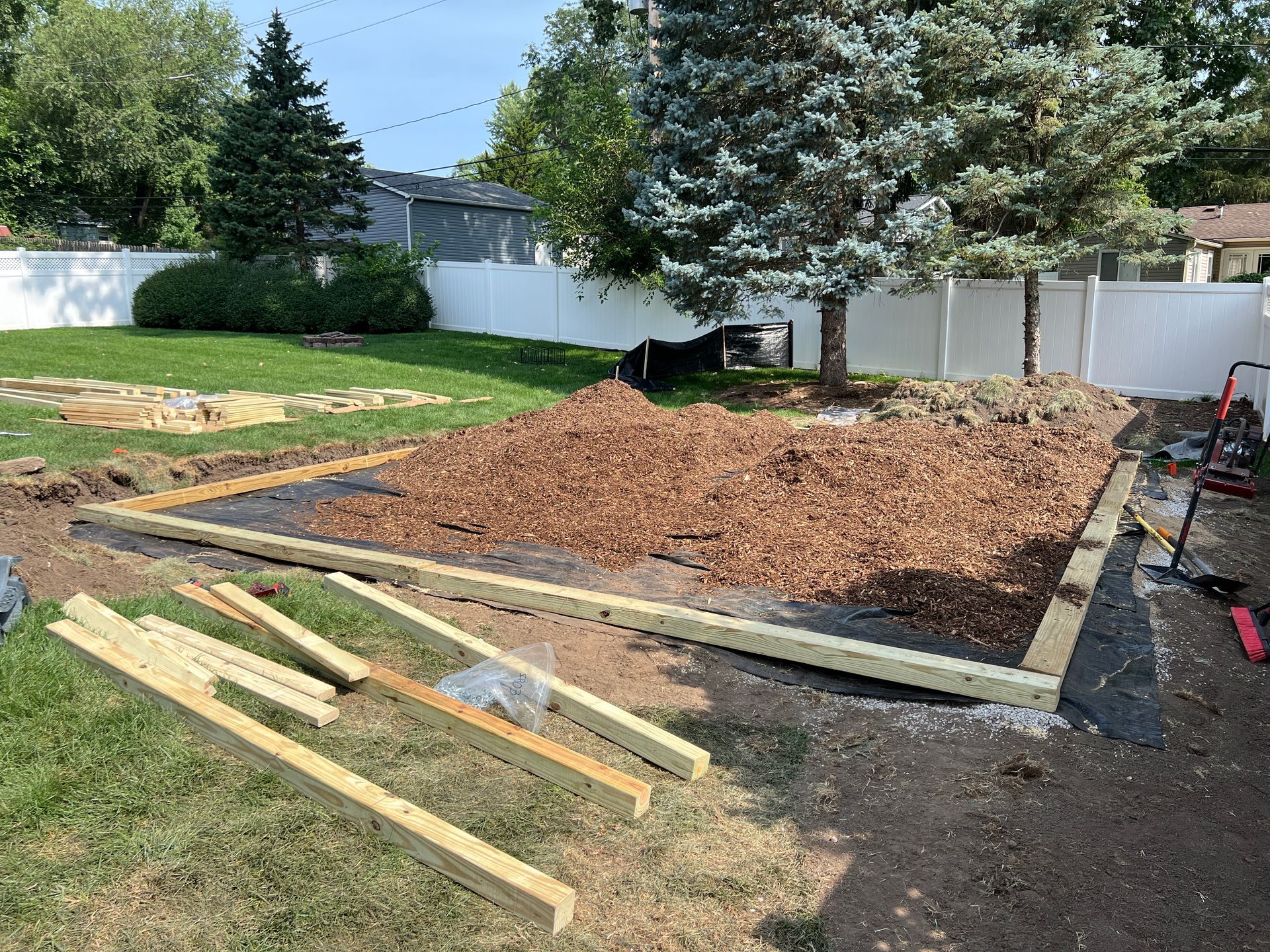 A backyard landscape project showing a framed rectangular base filled with mulch, with spare lumber in the foreground.