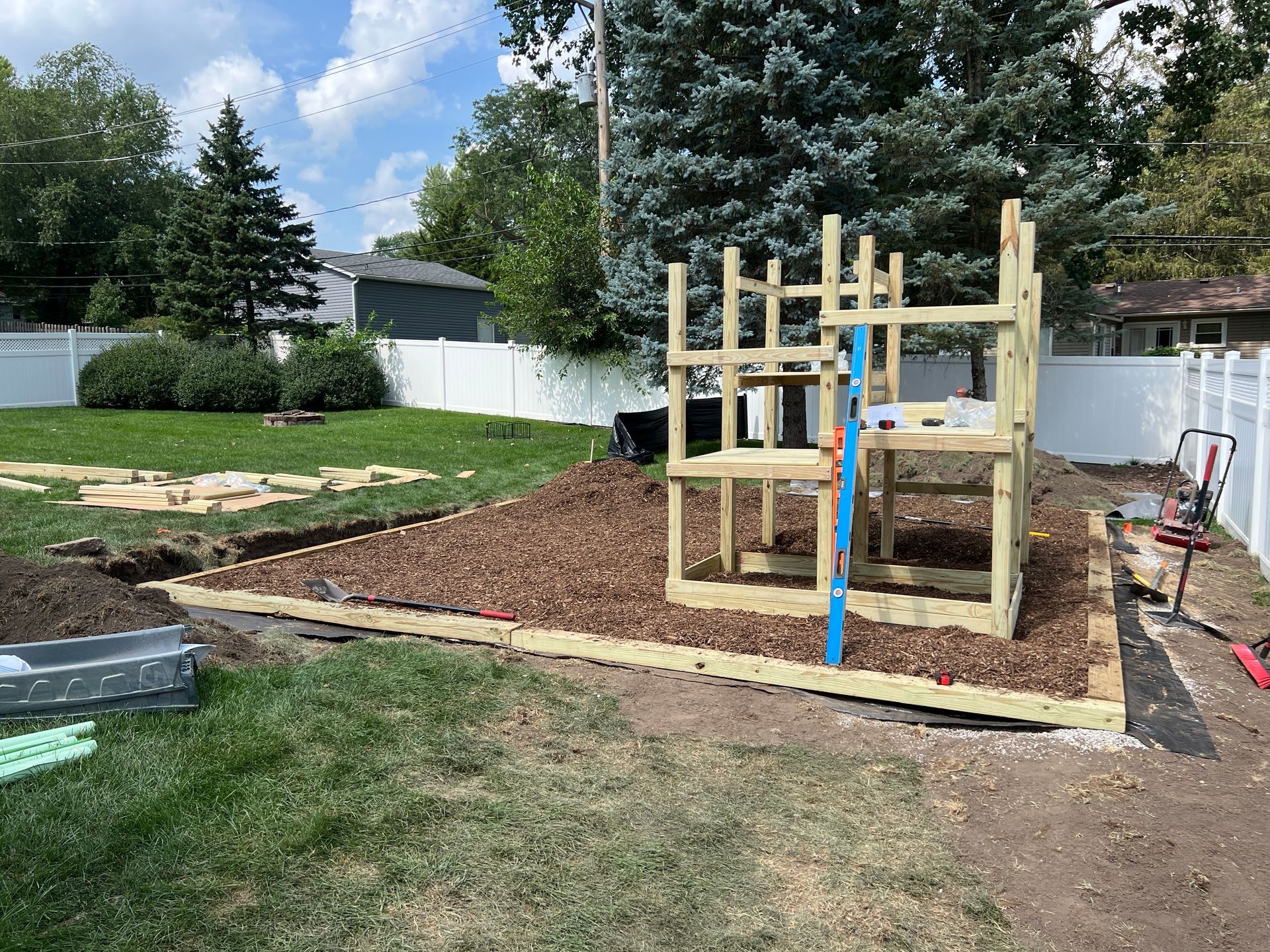 A wooden play structure under construction on a wood-mulched backyard site with a white fence in the background.