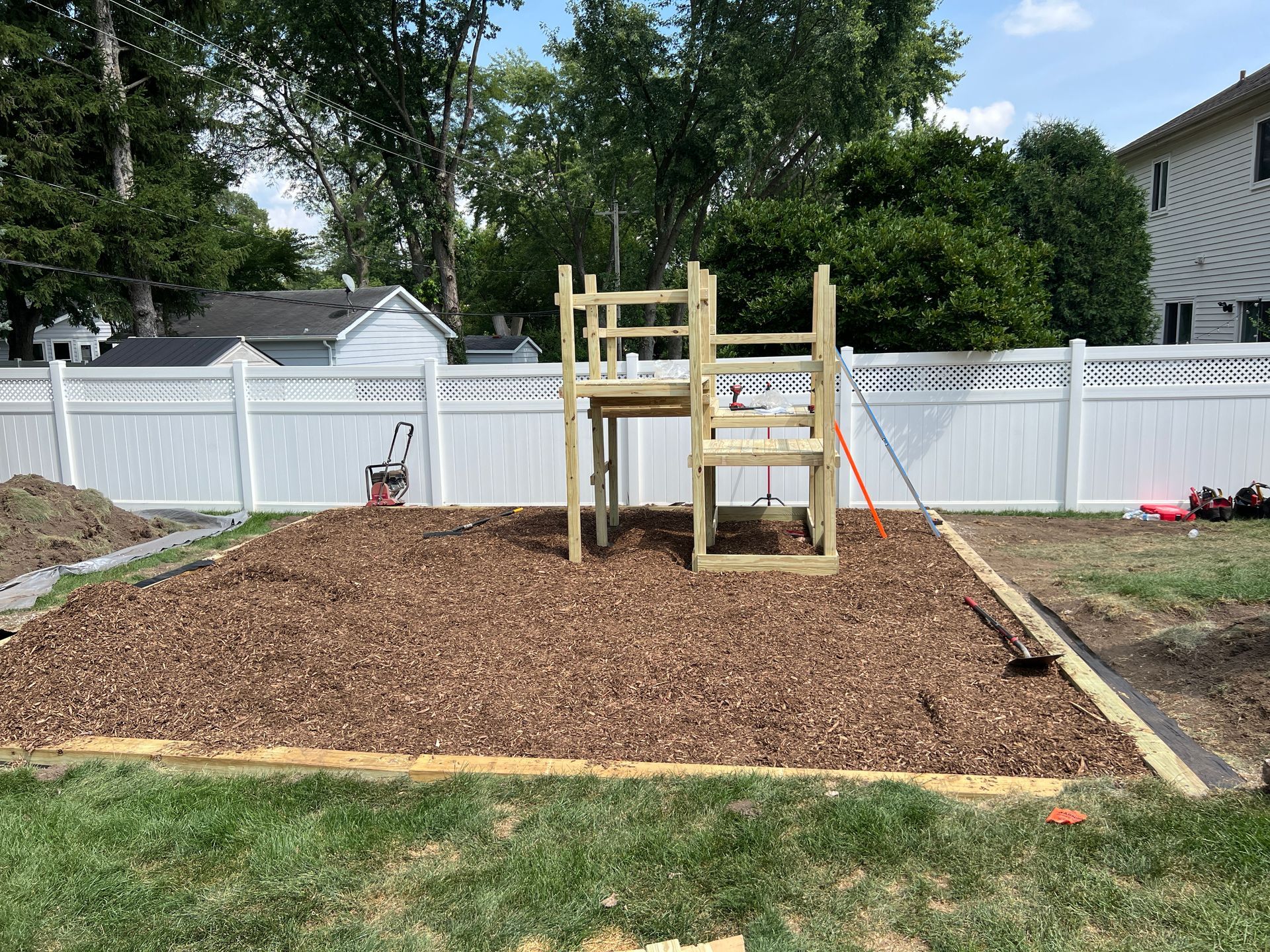 A wooden playset frame stands on a bed of dark mulch in a backyard, enclosed by a white fence.