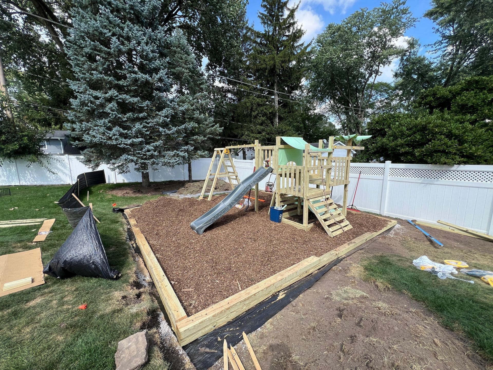 A newly constructed wooden playset with a green slide sits on a bed of wood mulch inside a rectangular landscape border.