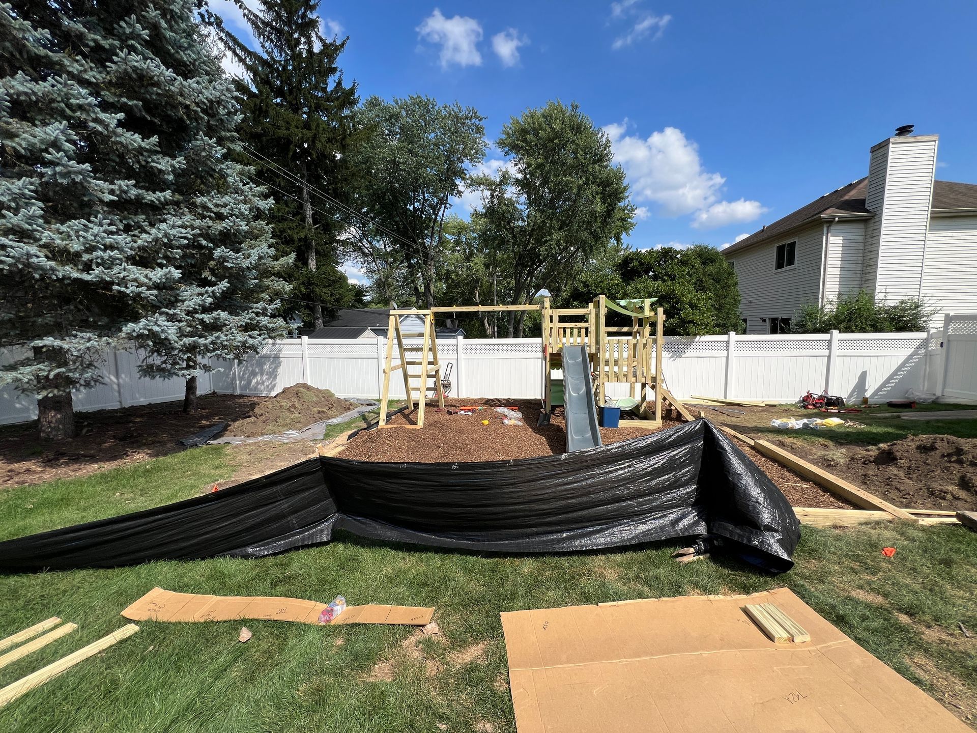 A backyard under construction with a wooden playset, a black landscape fabric barrier, and cardboard on the grass.