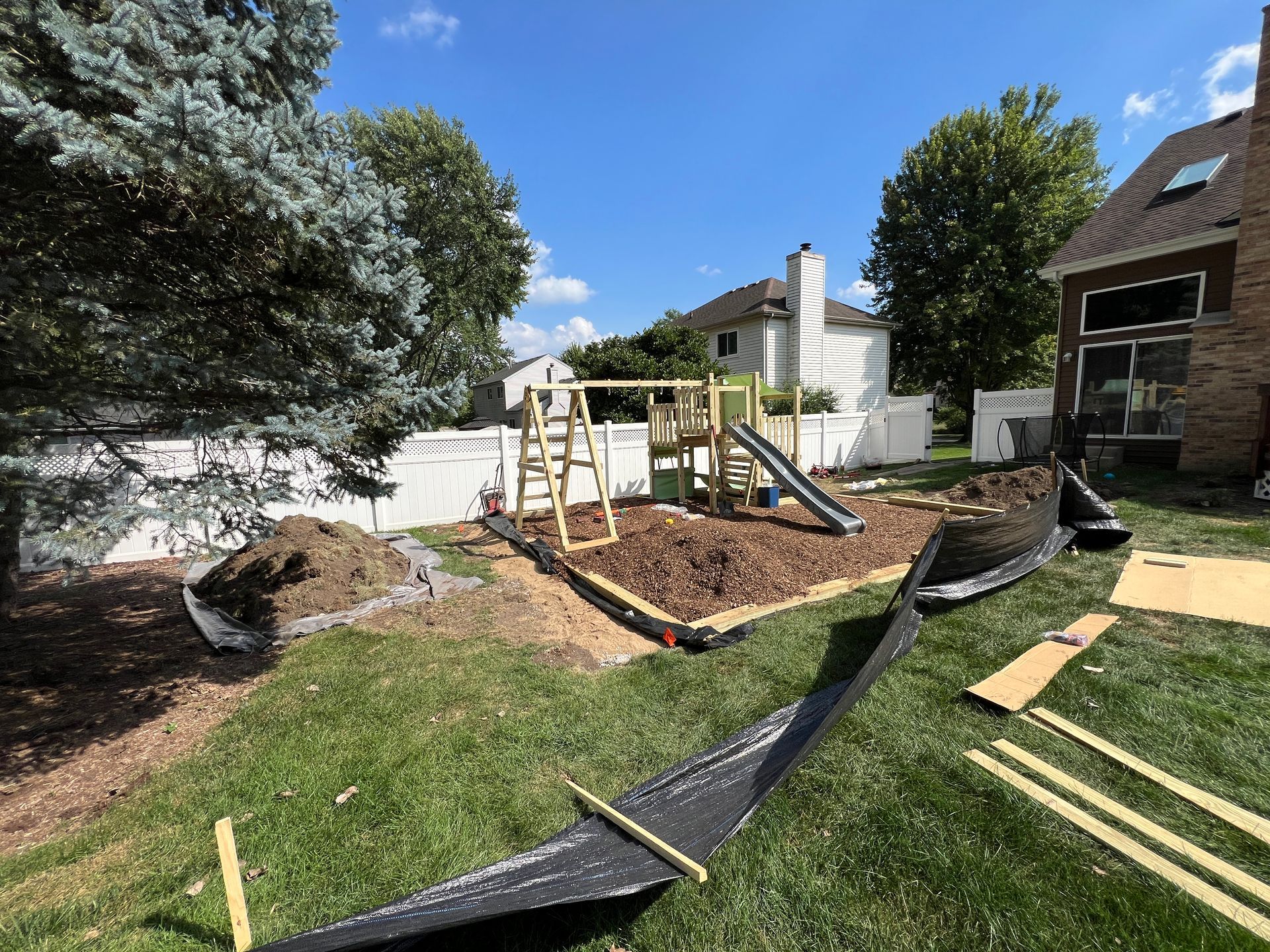 A backyard play set under construction with a wooden frame and metal slide inside an area marked by black landscaping film.