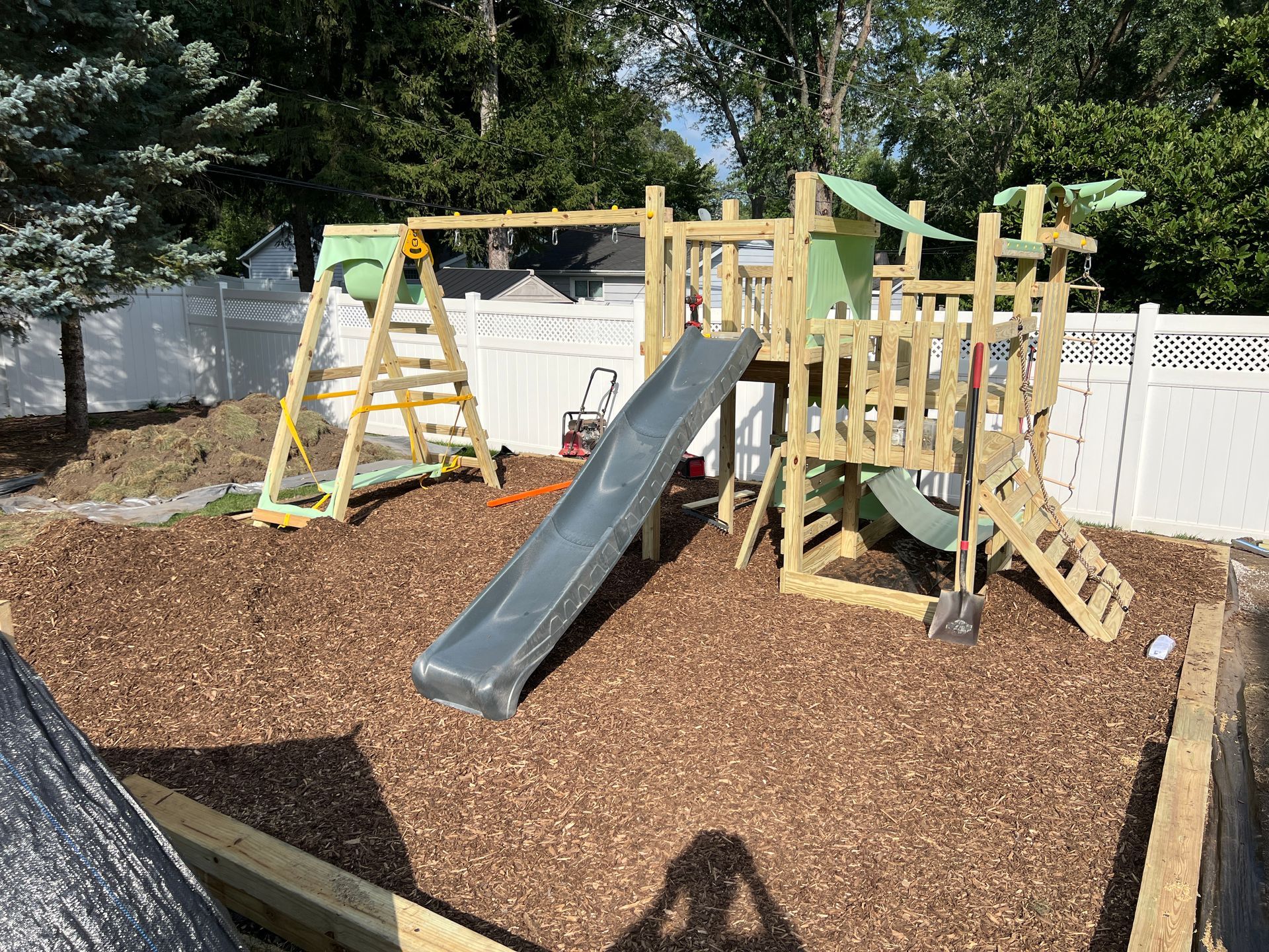A wooden backyard playset with a gray slide and a green canopy, situated in a mulch-covered area against a white fence.
