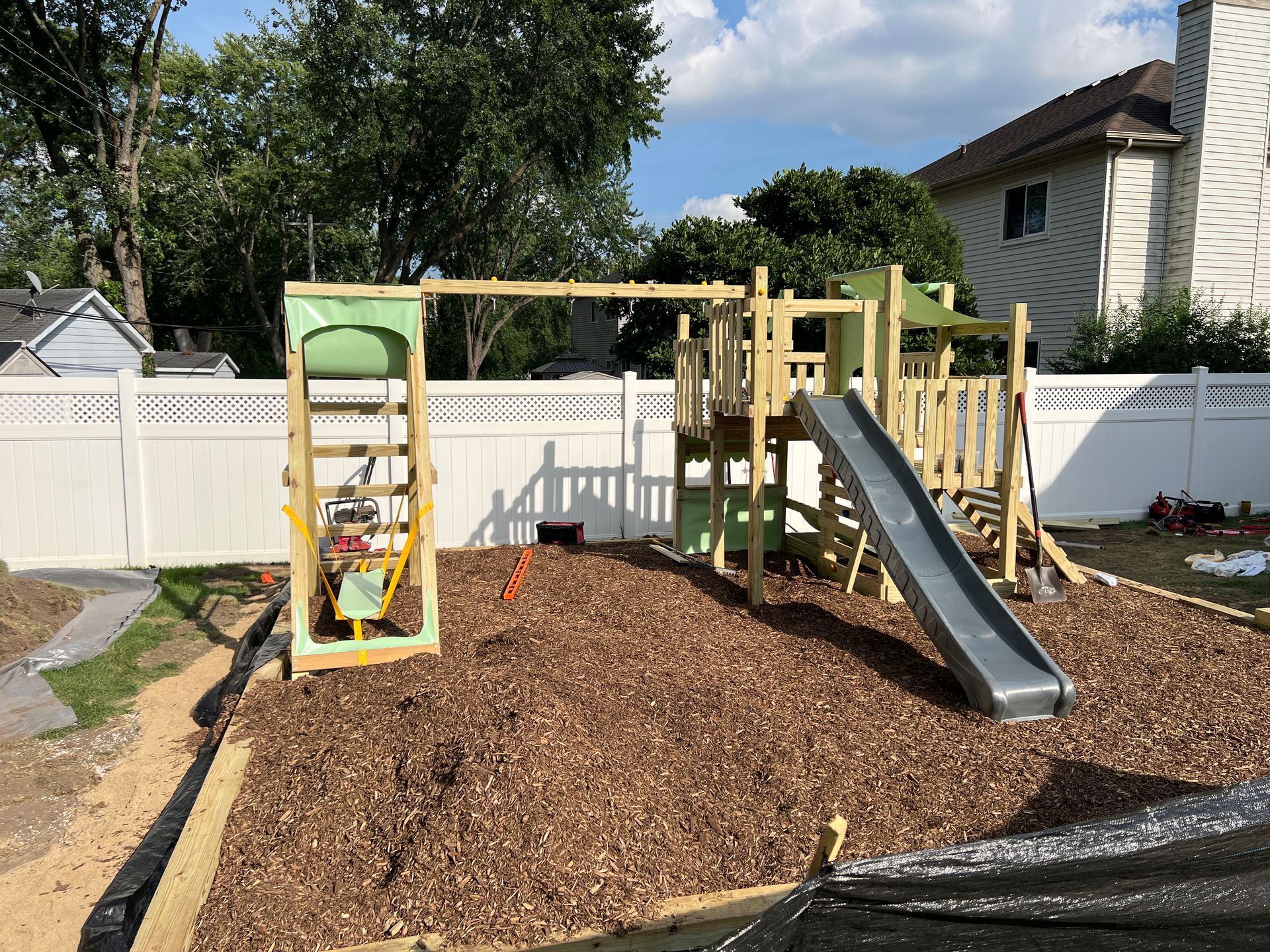 A wooden playground set with a slide and swings sits in a backyard on a wood-mulch surface in front of a white fence.