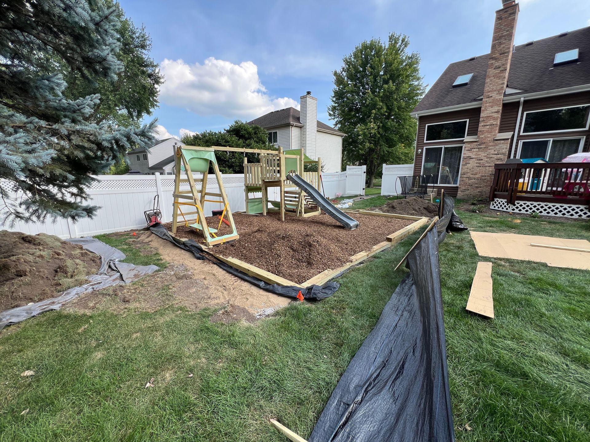 A wooden playset with a green slide sits on a bed of wood chips in a backyard, partially enclosed by black landscaping fabric.