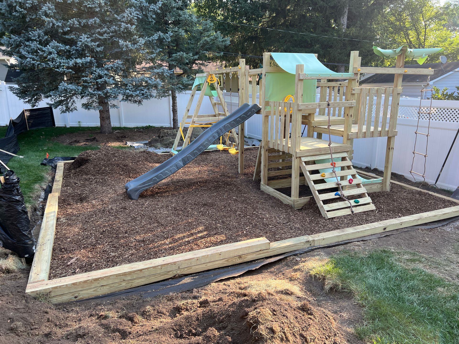 Wooden playset with slide and climbing wall set in a woodchip-filled area enclosed by landscape timbers in a backyard.