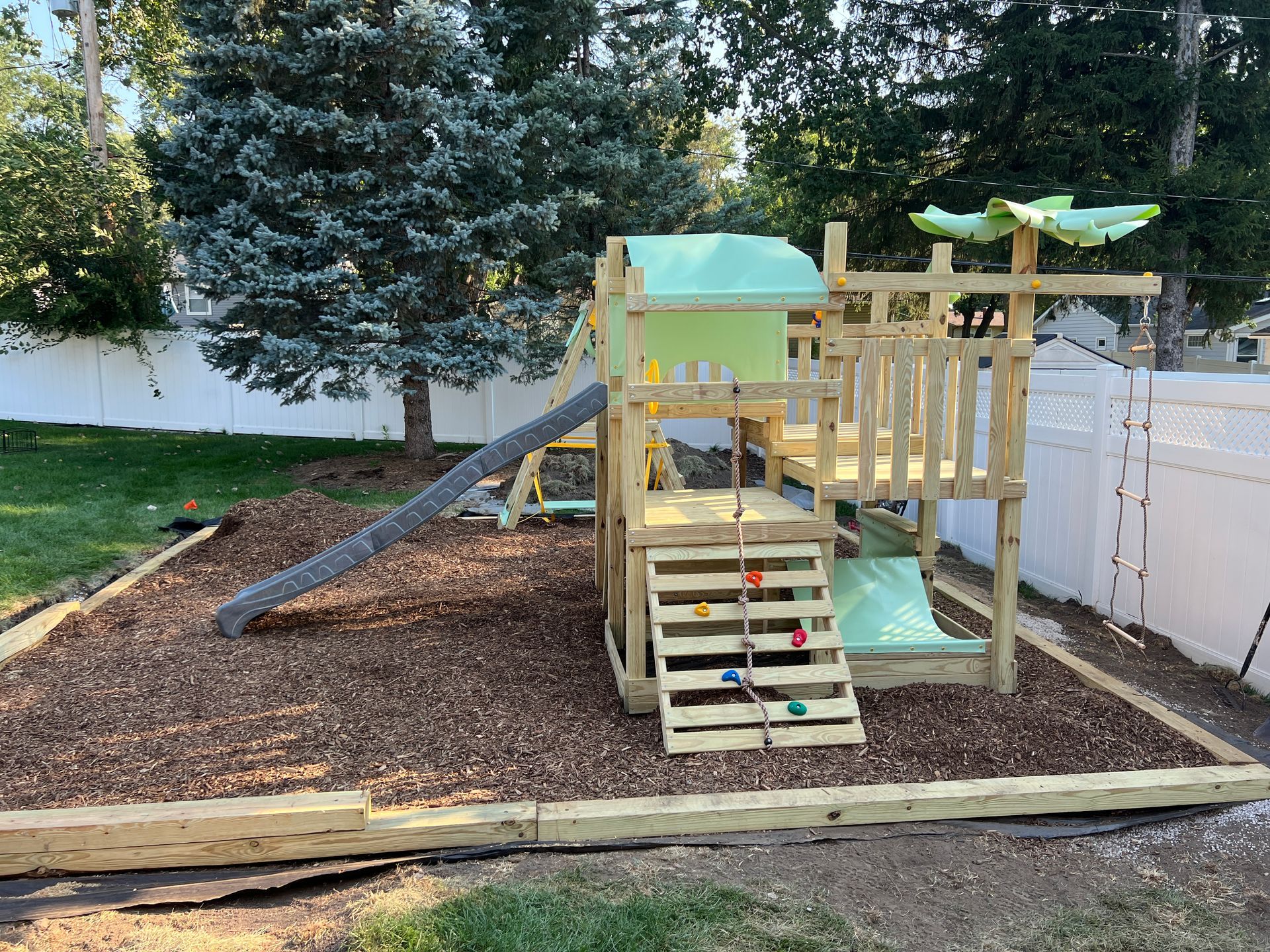 Wooden backyard playset with a climbing wall, slide, and canopy set on a wood-mulched area in a residential yard.