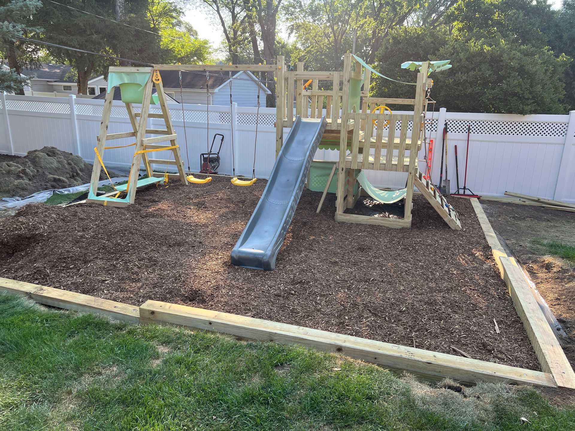 A wooden backyard swing set with a slide and a rope ladder, situated in a mulch-covered area bordered by timber beams.