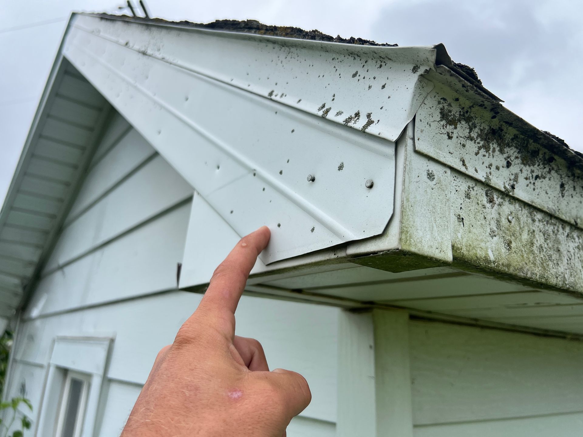 A person's finger points to the white metal trim on the edge of a house roof, showing signs of wear and peeling paint.