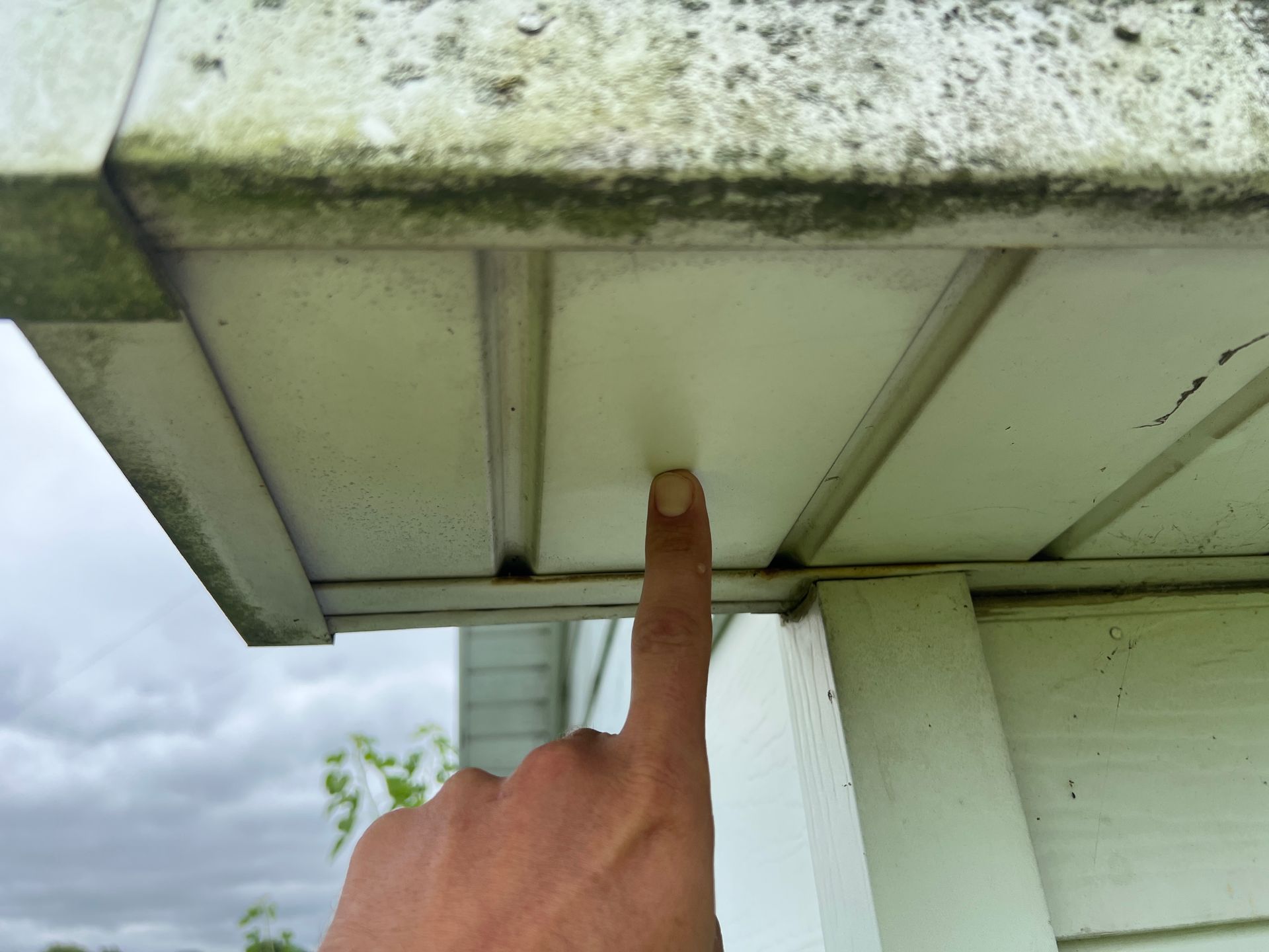 A finger points to an area of discolored, weathered white siding under the edge of a roof eaves.