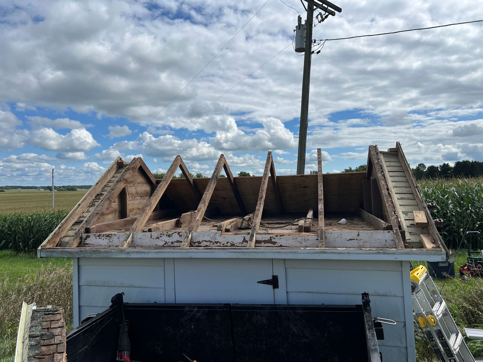 A shed with the roof shingles removed, exposing the rafters and plywood sheathing, set in a rural field under a blue sky.