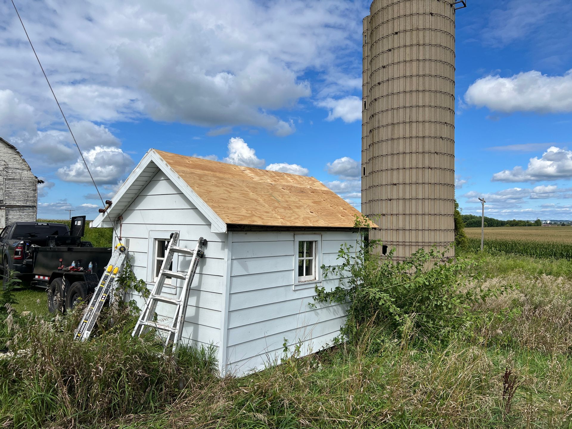 A small white shed with a newly shingled roof stands beside a tall cylindrical silo under a blue, cloudy sky.