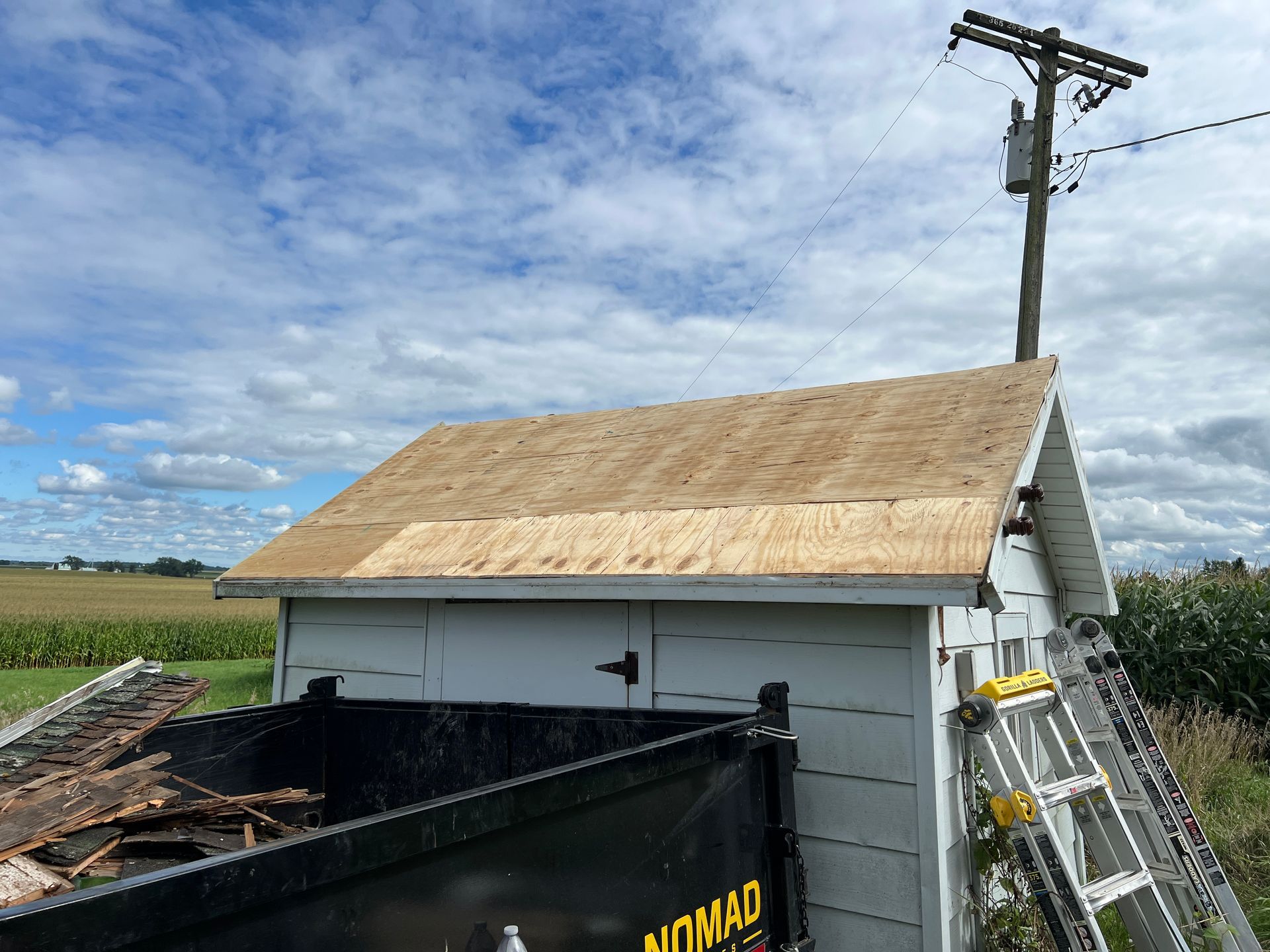 A white shed with a freshly installed plywood roof stands next to a dumpster under a utility pole on a cloudy day.