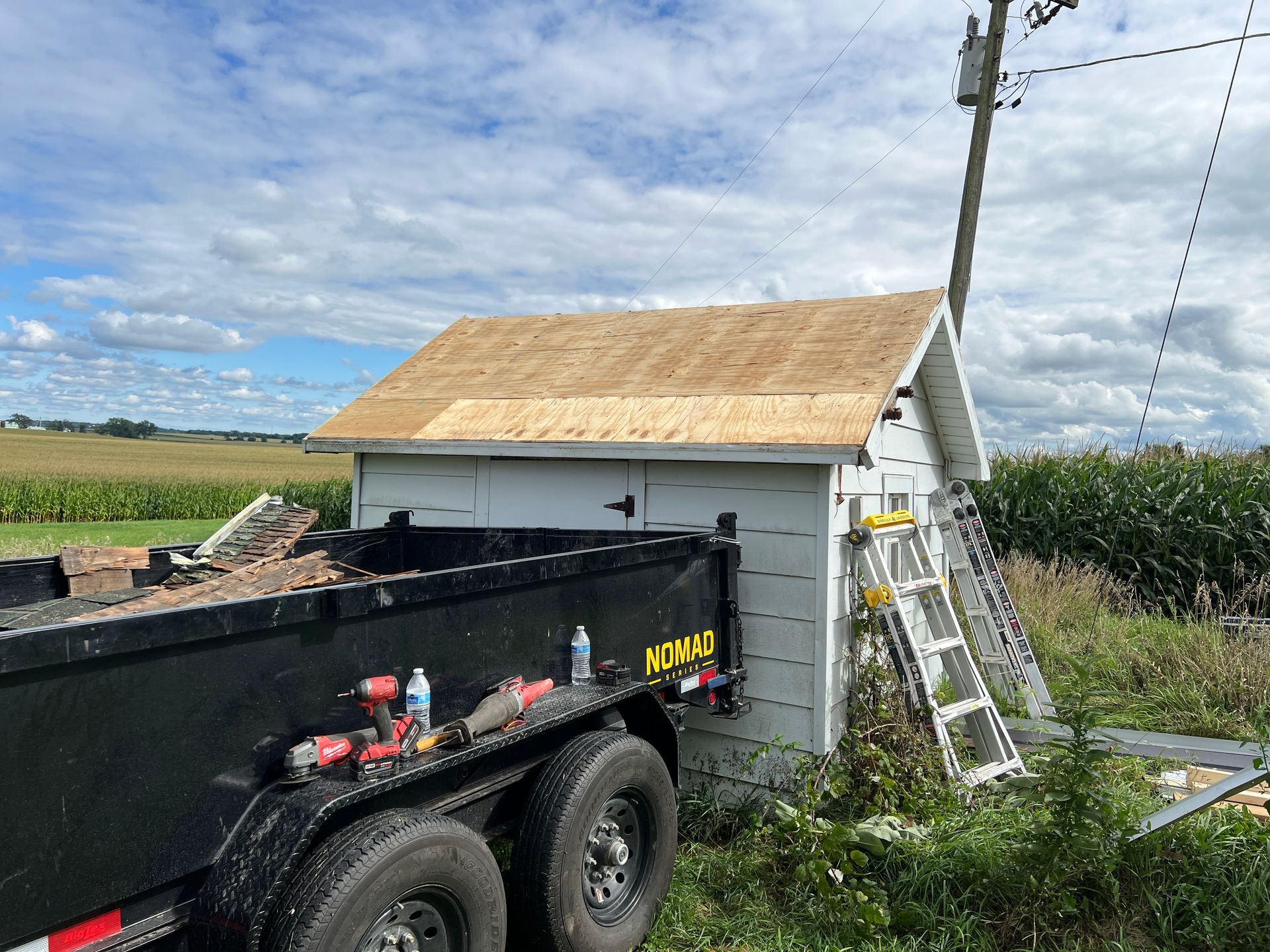 A black dump trailer sits beside a small, white shed with a newly sheathed plywood roof under a cloudy blue sky.