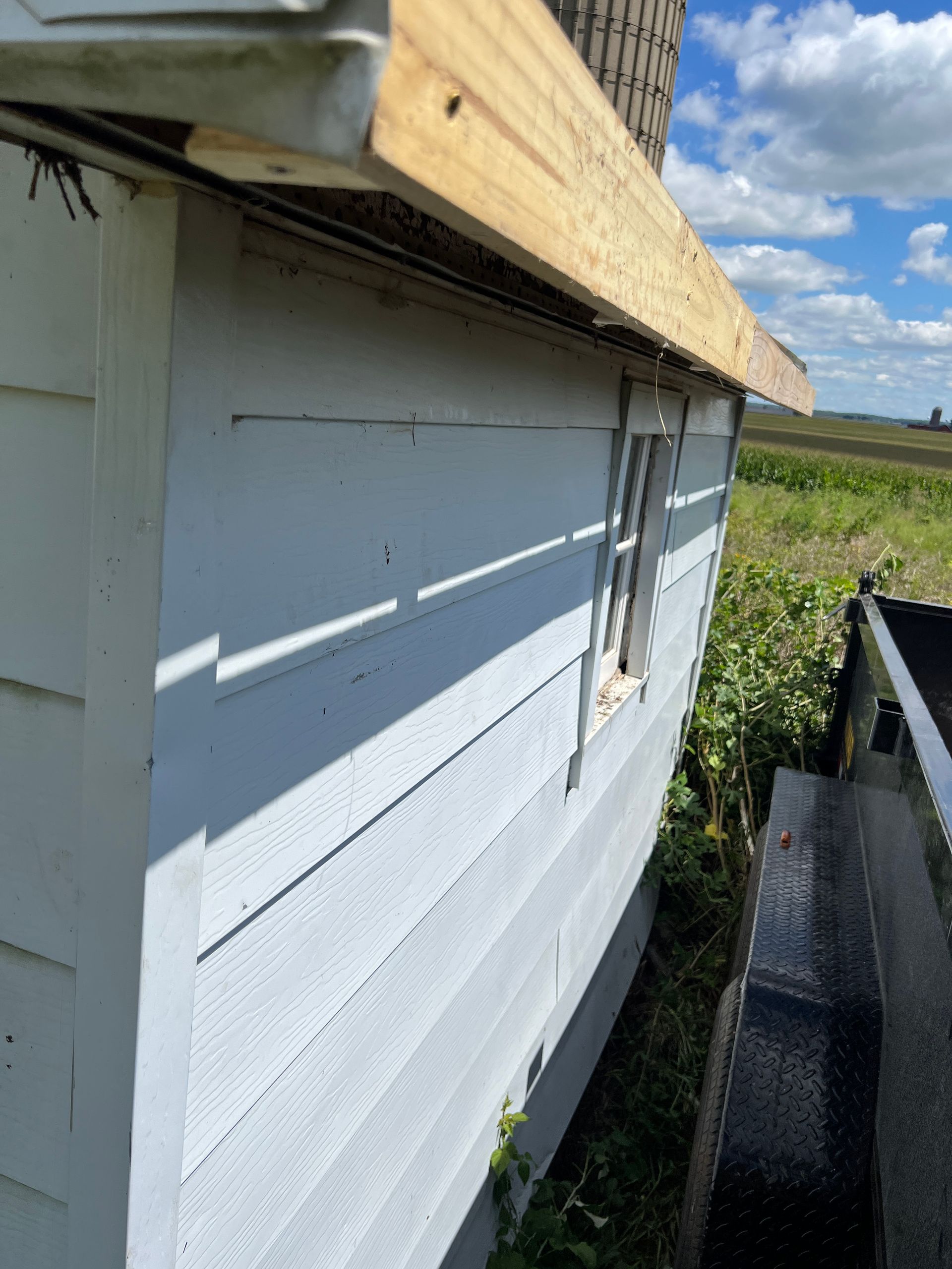 A side view of a white, lap-sided shed with a new wooden trim board under the roof edge, set in a rural field.