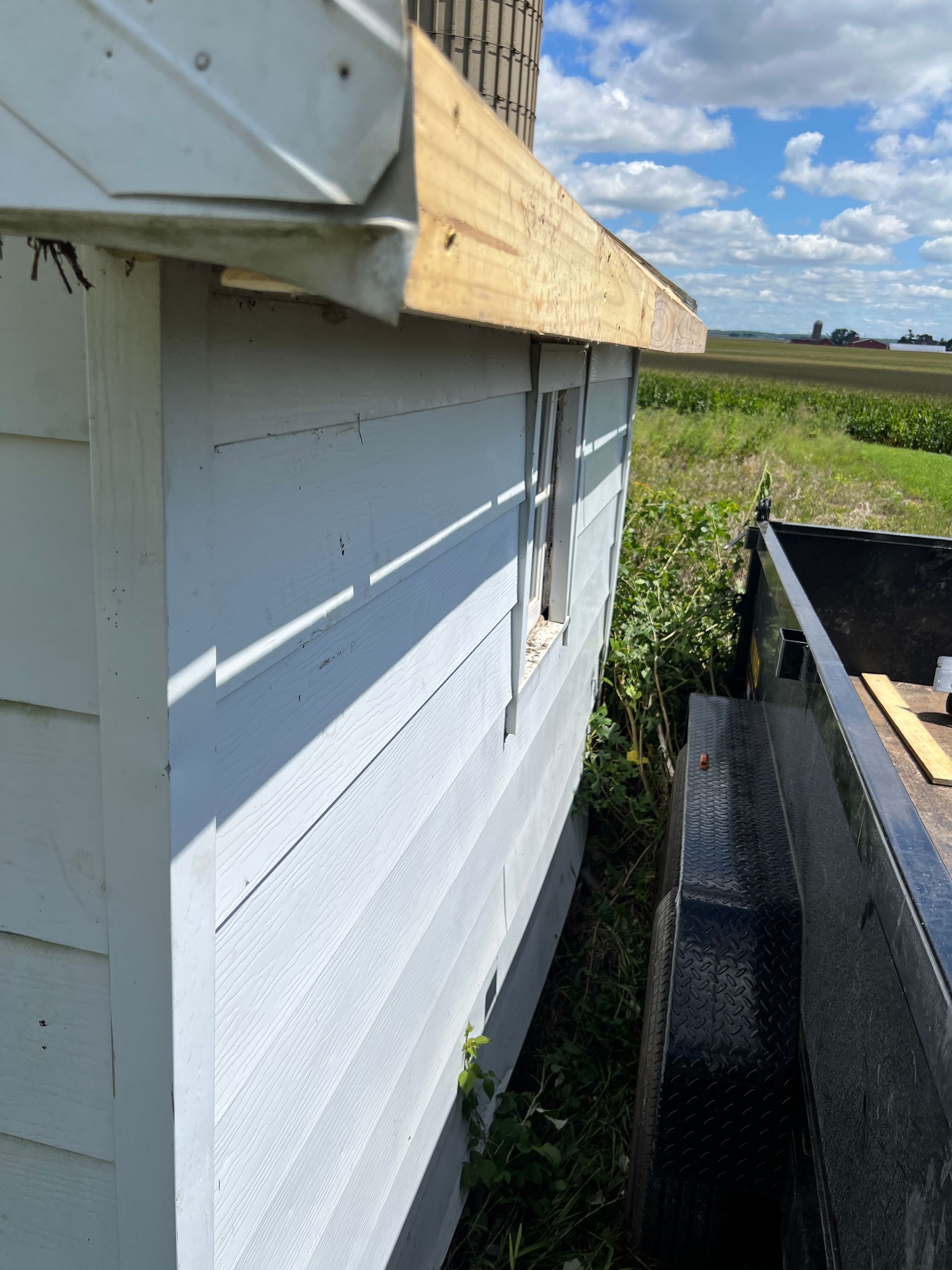 A light blue shed exterior featuring a newly installed wooden fascia board, viewed from beside a dark utility trailer.