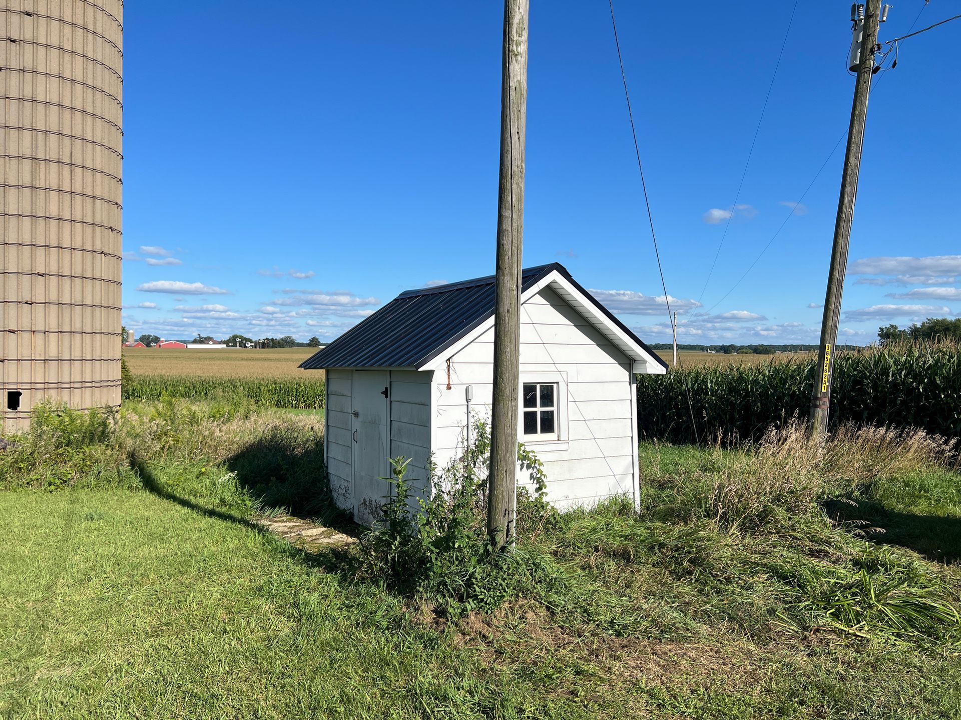 A white shed with a dark roof stands in a grassy field next to a tall silo and a utility pole under a blue sky.