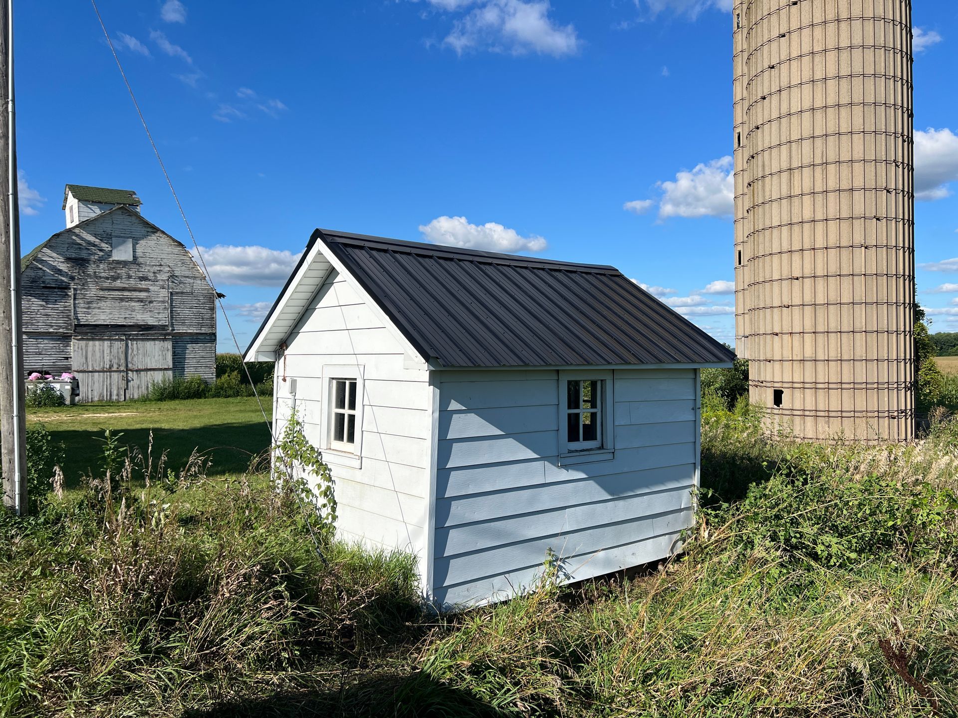 A small white shed with a black roof sits in a grassy field, flanked by a weathered barn and a tall silo under a blue sky.