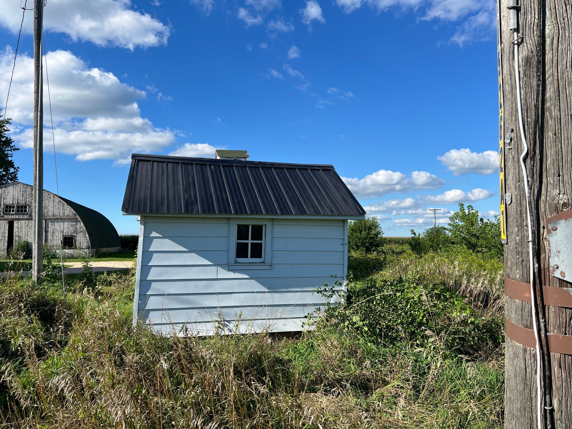 A small, white wooden shed with a black metal roof sits in a field under a blue sky with scattered clouds.