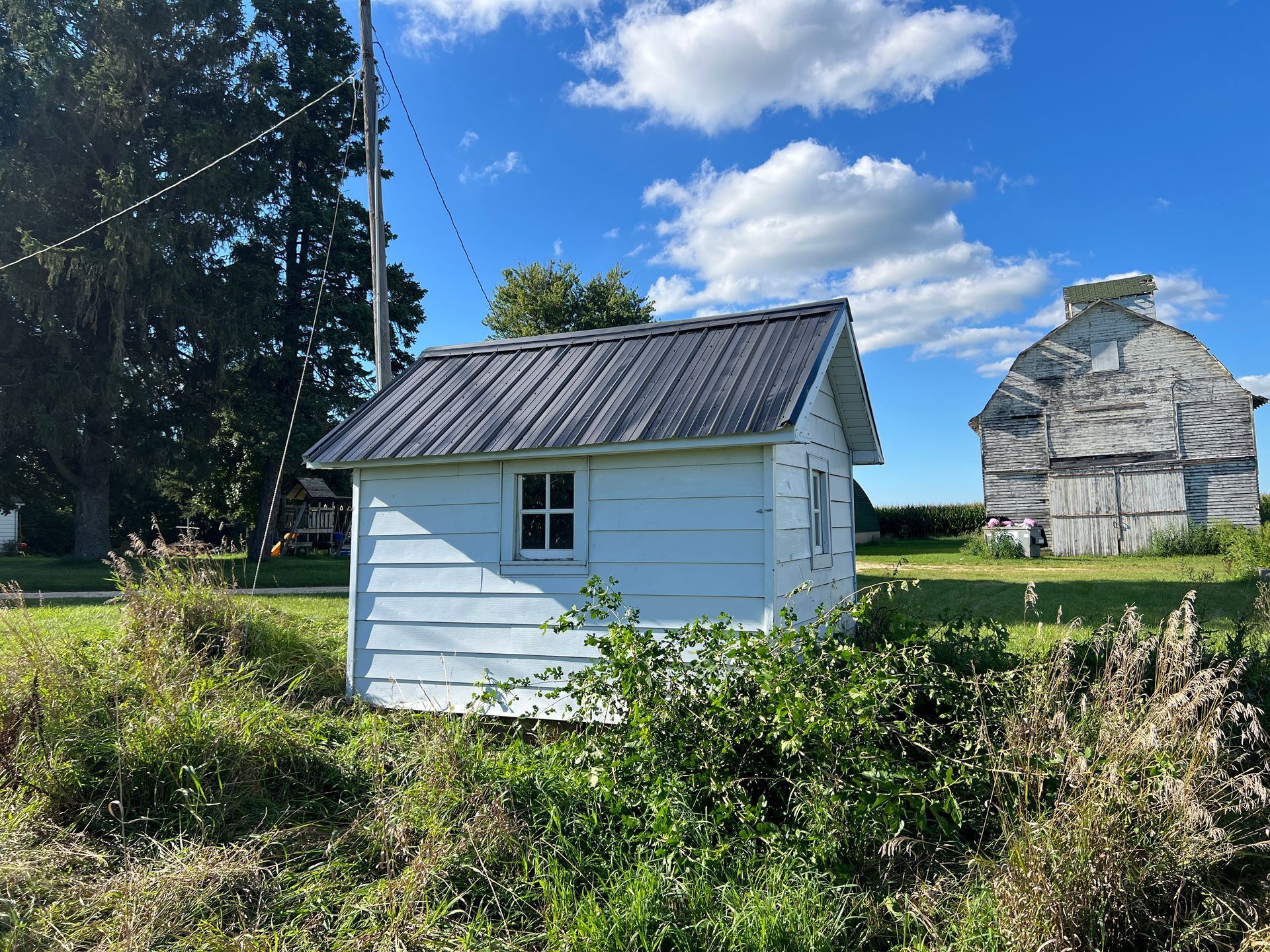 A small white shed with a dark metal roof sits in a grassy field, with a large, weathered white barn in the background.