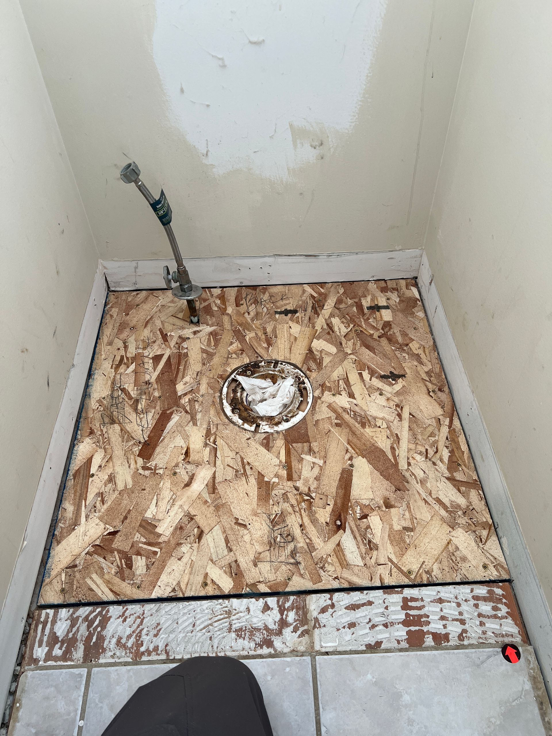 A bathroom floor with an exposed wooden subfloor, a toilet flange, and a water supply line near a tiled area.