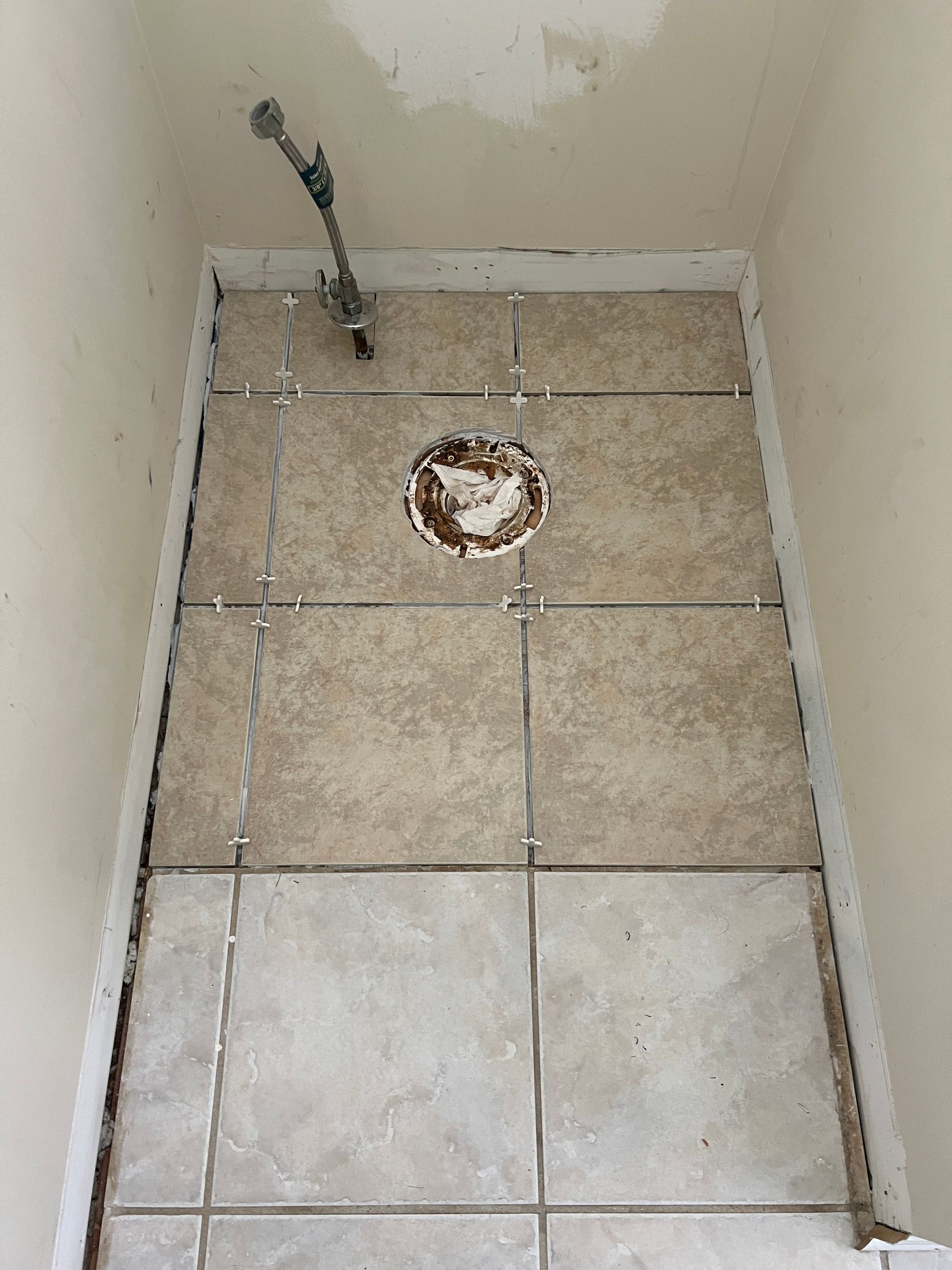 A partially tiled bathroom floor with a toilet flange in the center and a water supply line in the corner.