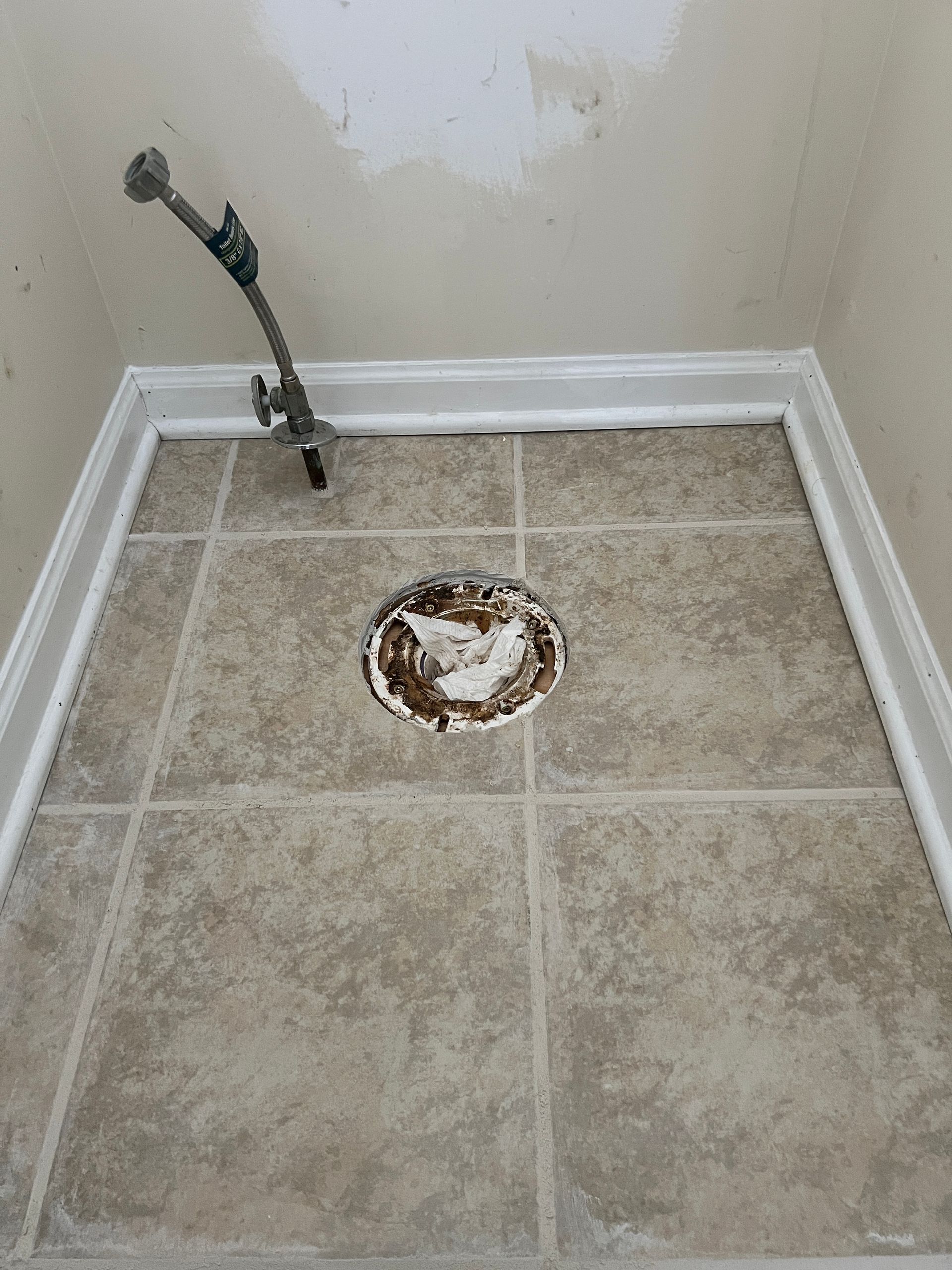 A floor view of a bathroom showing a circular drain opening in tan tiled flooring and a water supply line in the corner.