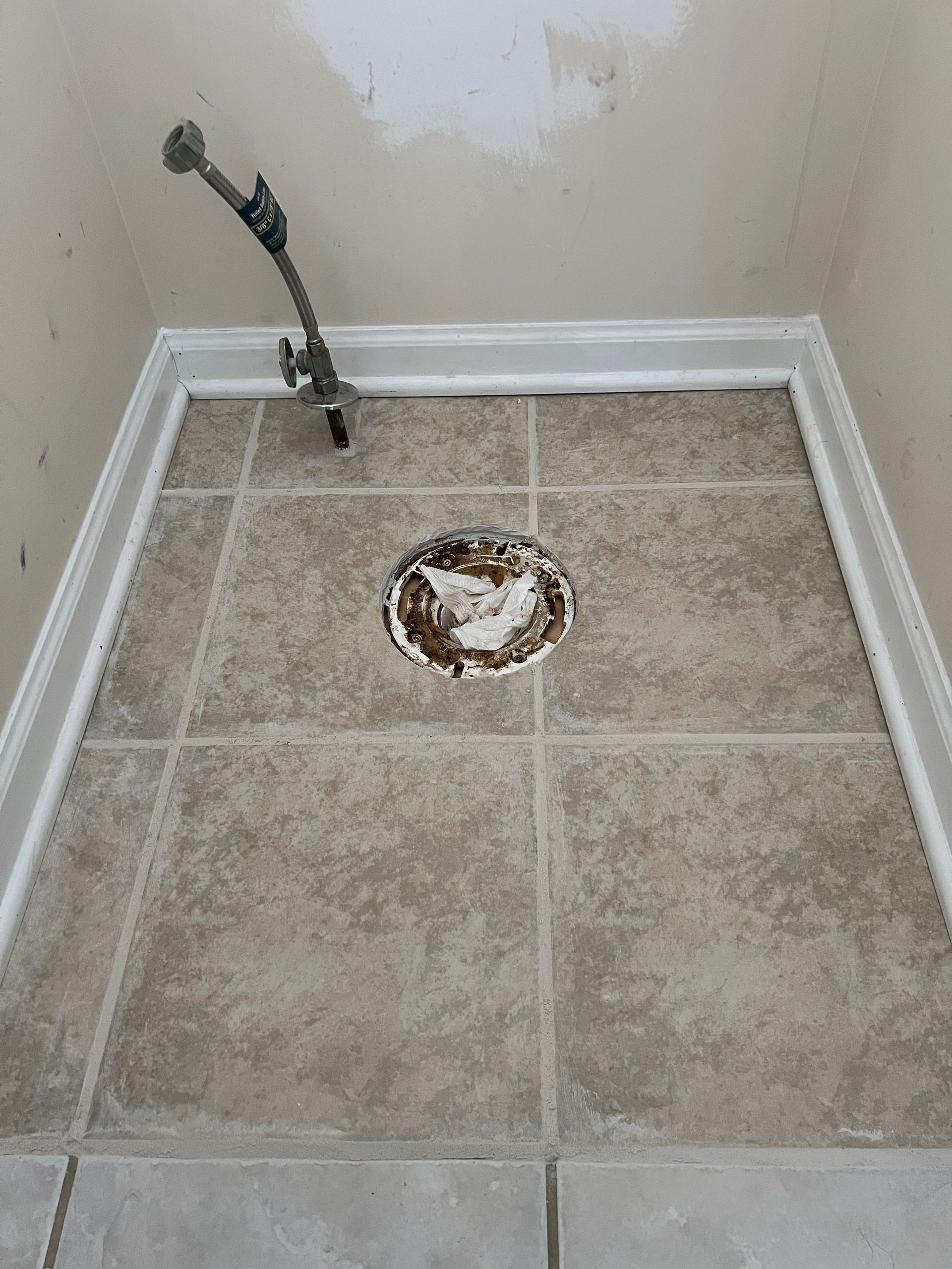A bathroom floor with tan square tiles, a central toilet drain opening, and a water supply line emerging from the wall.