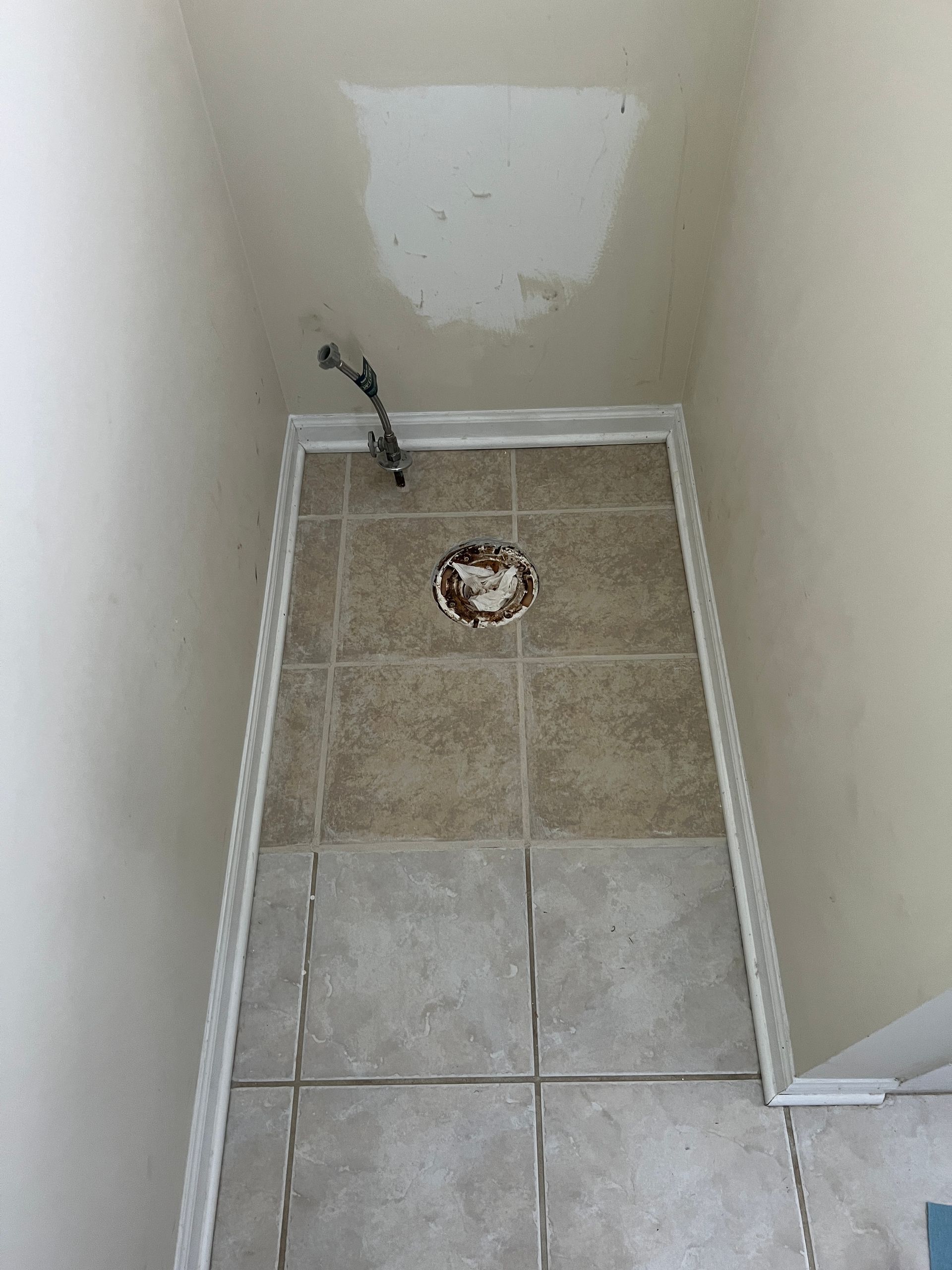 A high-angle view of a small bathroom alcove showing a tiled floor, a toilet flange, and a water supply valve on the wall.