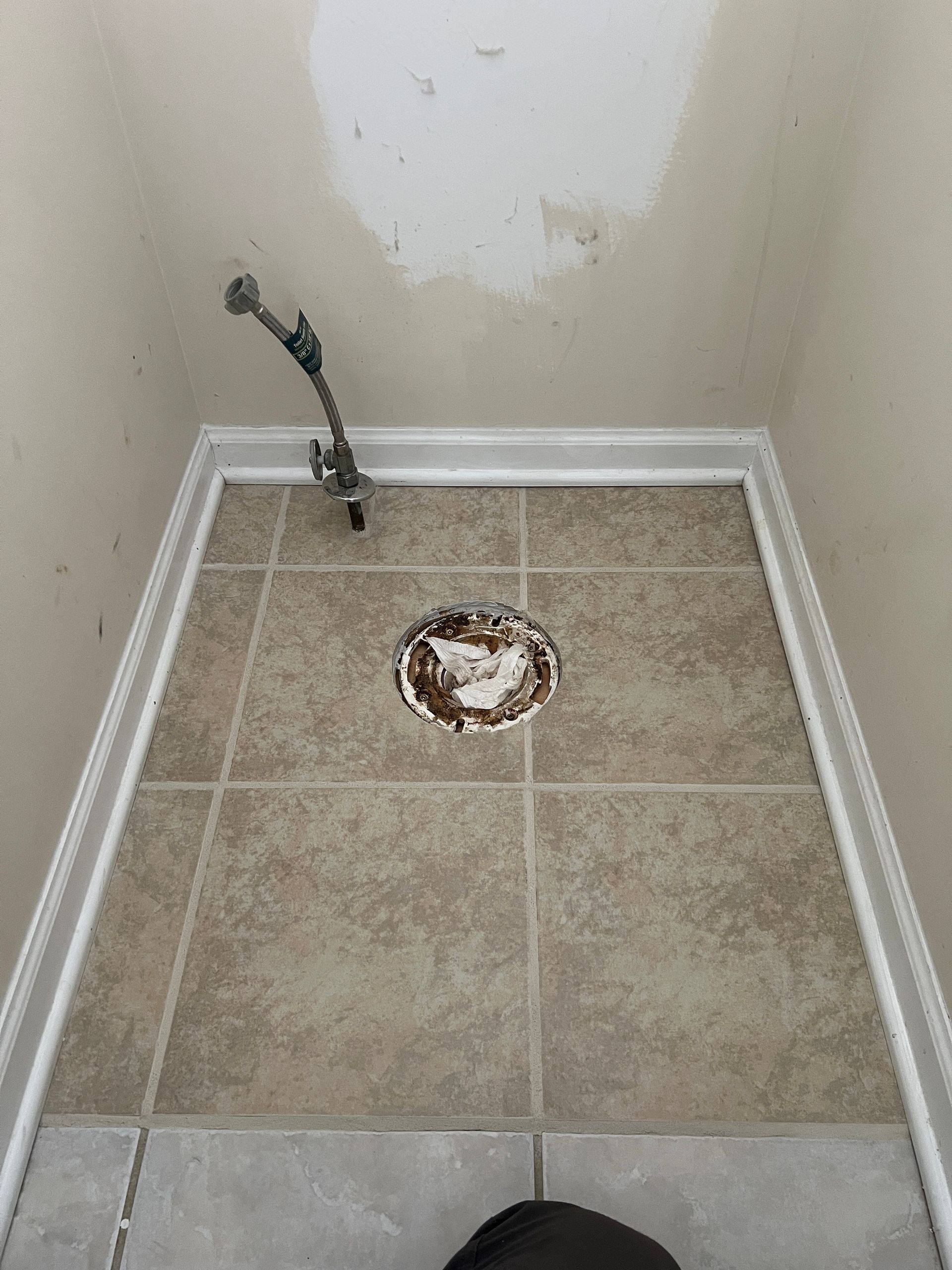 A residential bathroom floor with a tan tile pattern, a circular drain opening, and a water supply line near the wall.