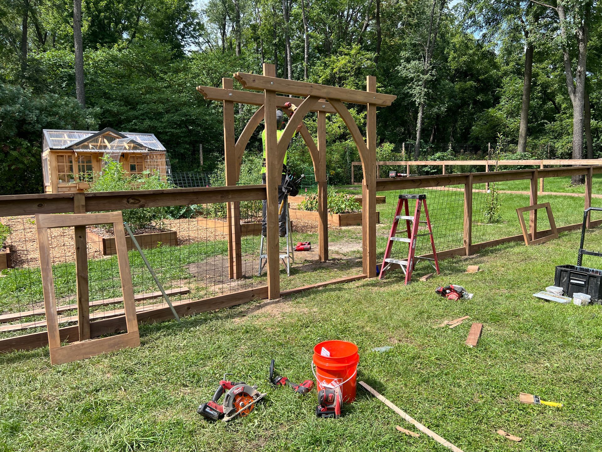 A wooden garden arbor and fence under construction in a grassy yard, with tools and a stepladder nearby.