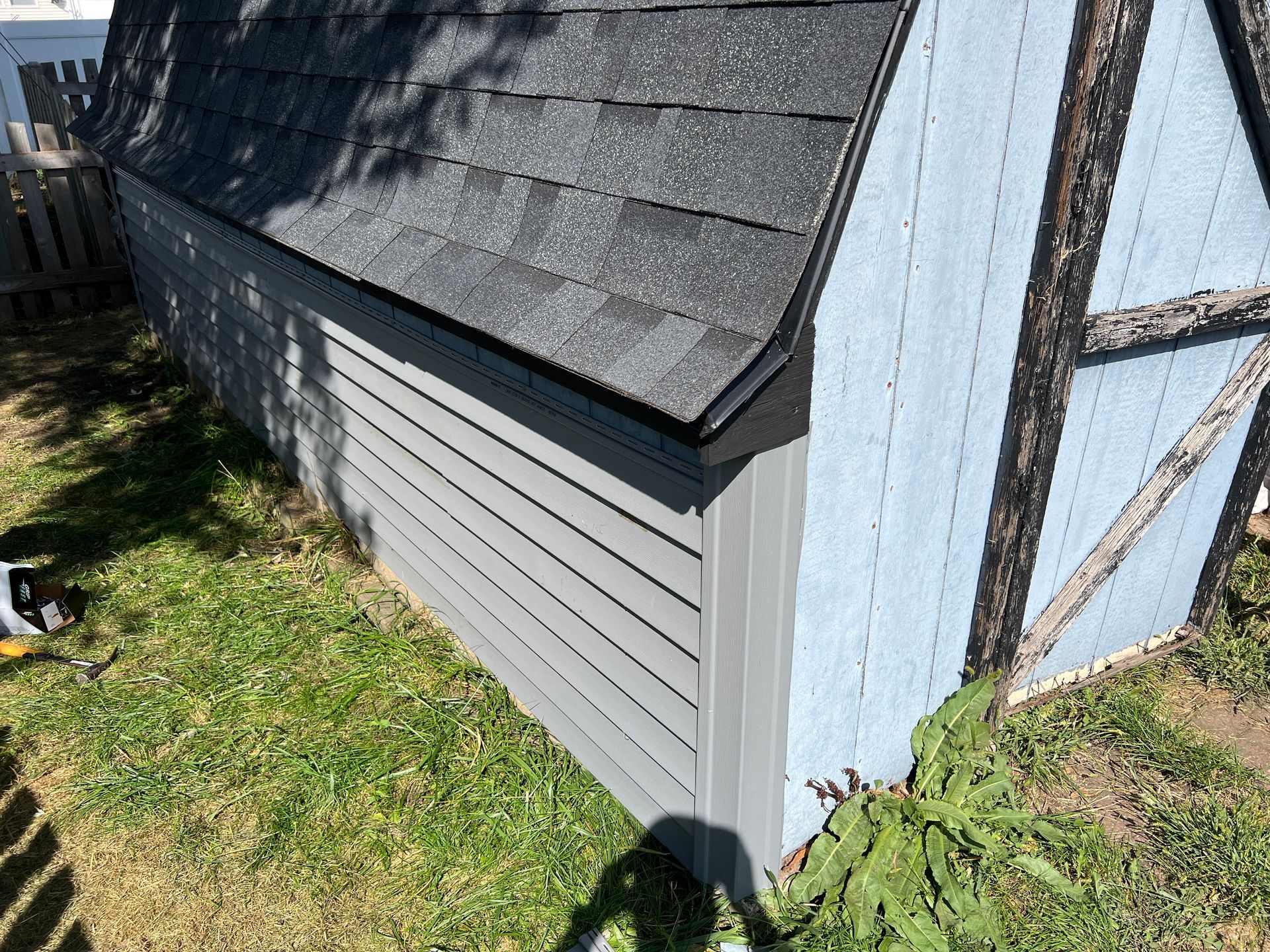 A low-angle view of a backyard shed with a grey shingled roof, light blue siding, and horizontal grey wall panels.