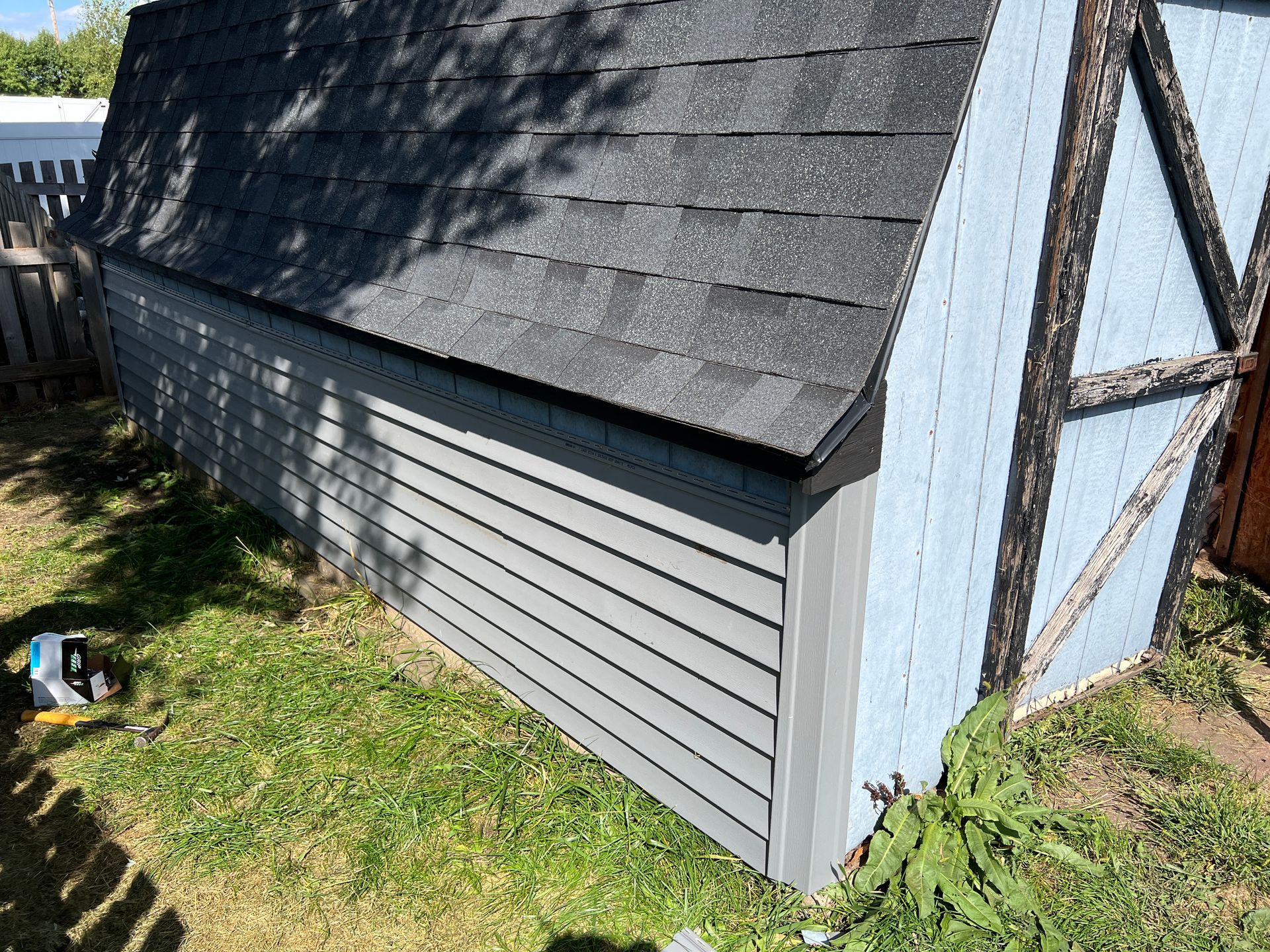 A small shed in a grassy yard, featuring light blue siding, a dark trim, and a dark shingled roof.