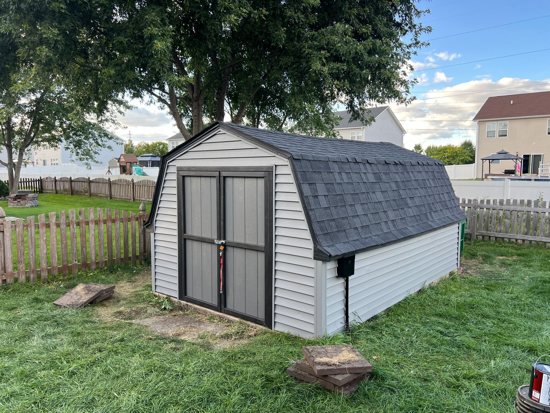A gray, vinyl-sided storage shed with a dark shingled gambrel roof sits in a grassy backyard near a wooden fence.