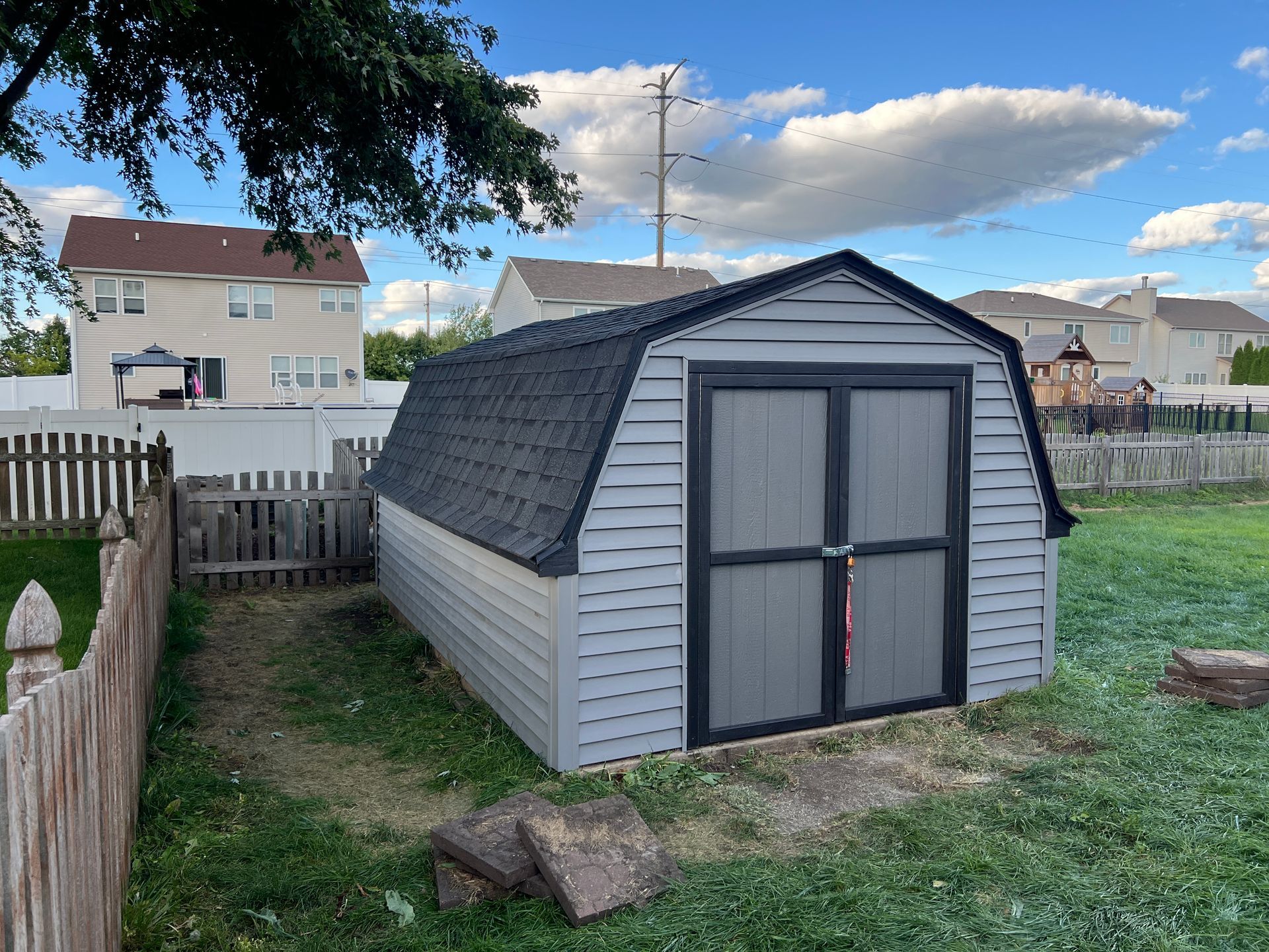 A gray storage shed with a dark roof sits on a grassy lawn in a residential backyard.