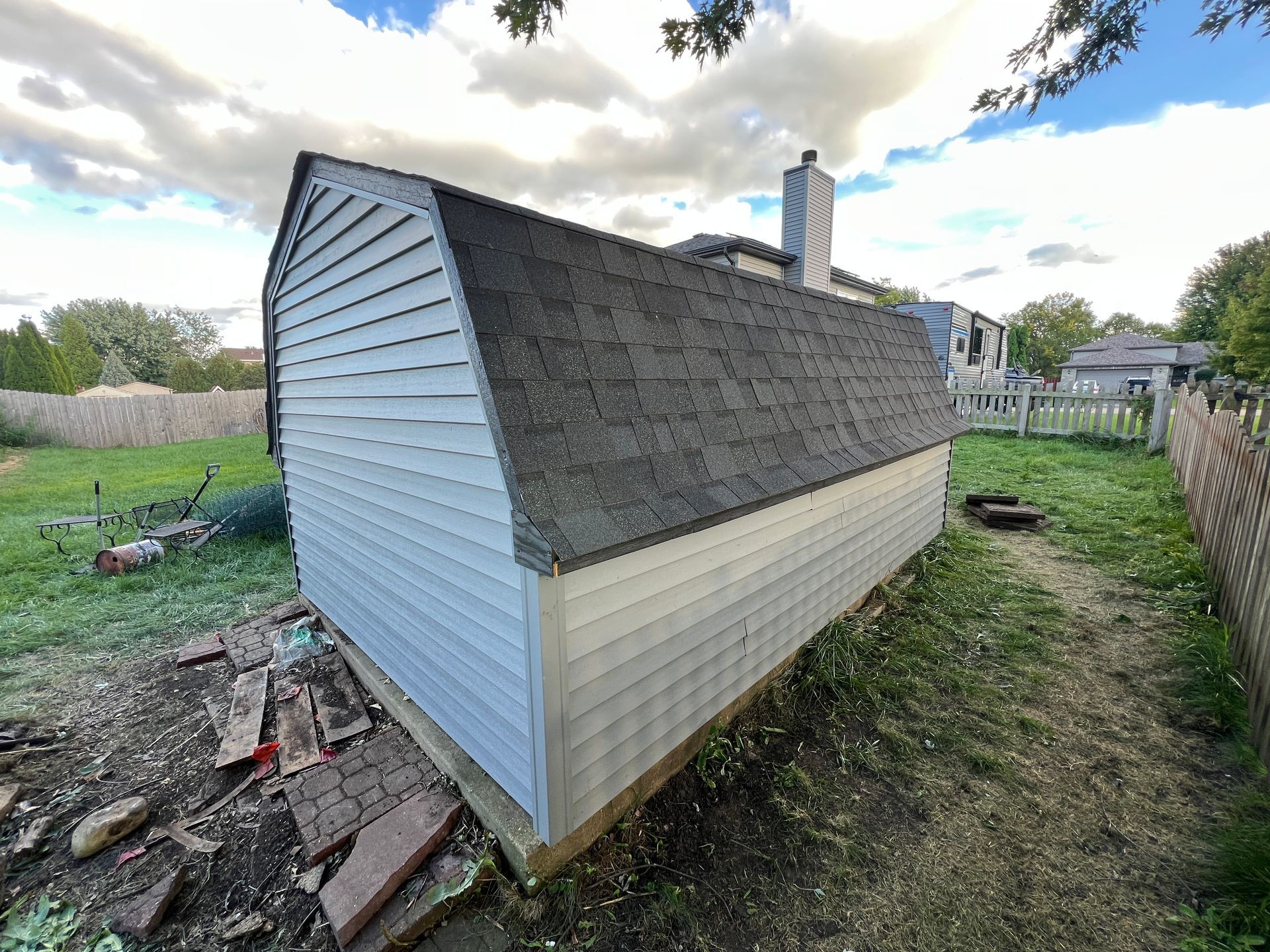 A light blue, gambrel-roofed shed sits in a grassy backyard with scattered bricks and a partial wooden fence.