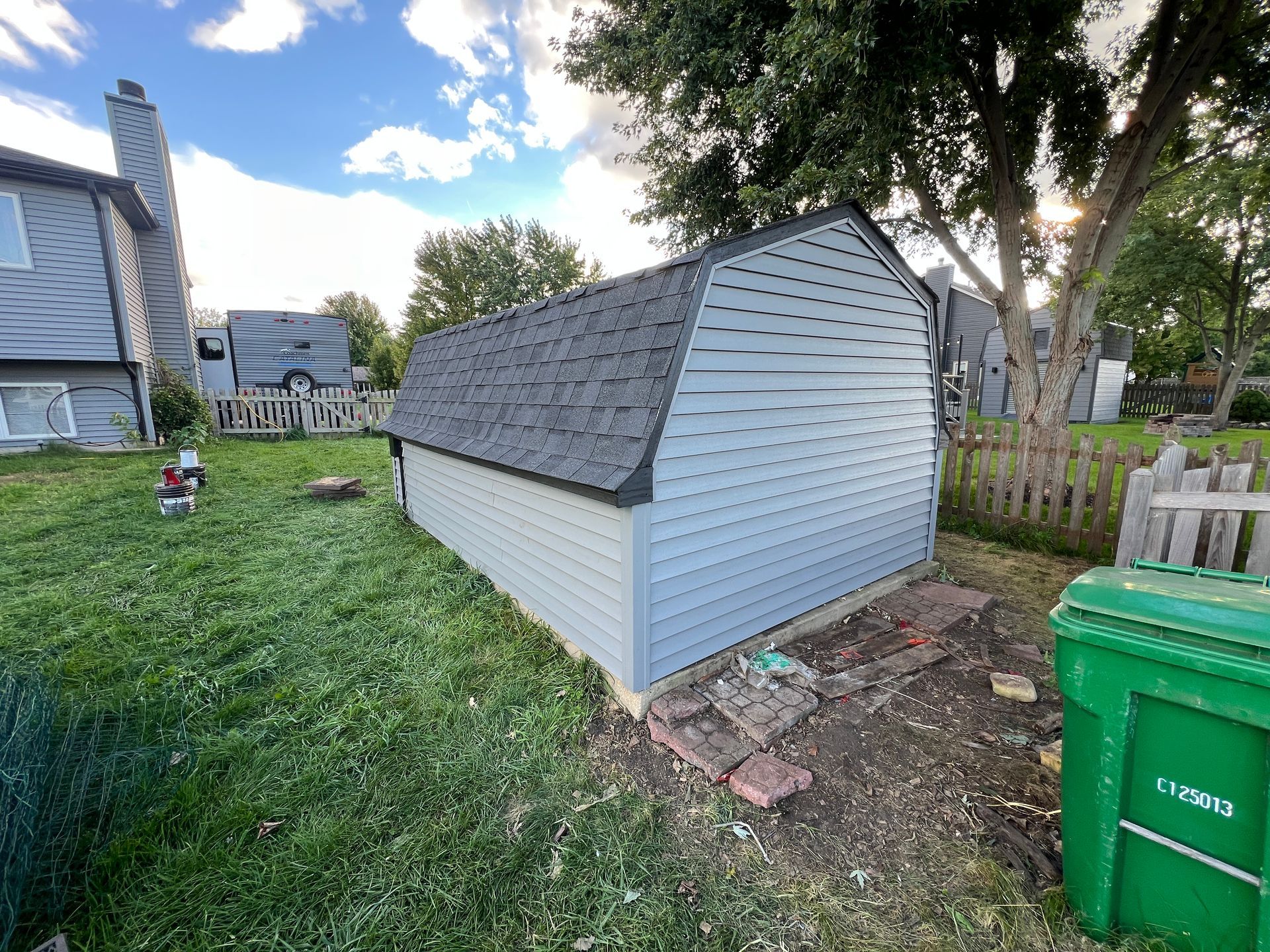A backyard shed with light gray vinyl siding and a dark shingled gambrel roof, set on a lawn next to a residential house.
