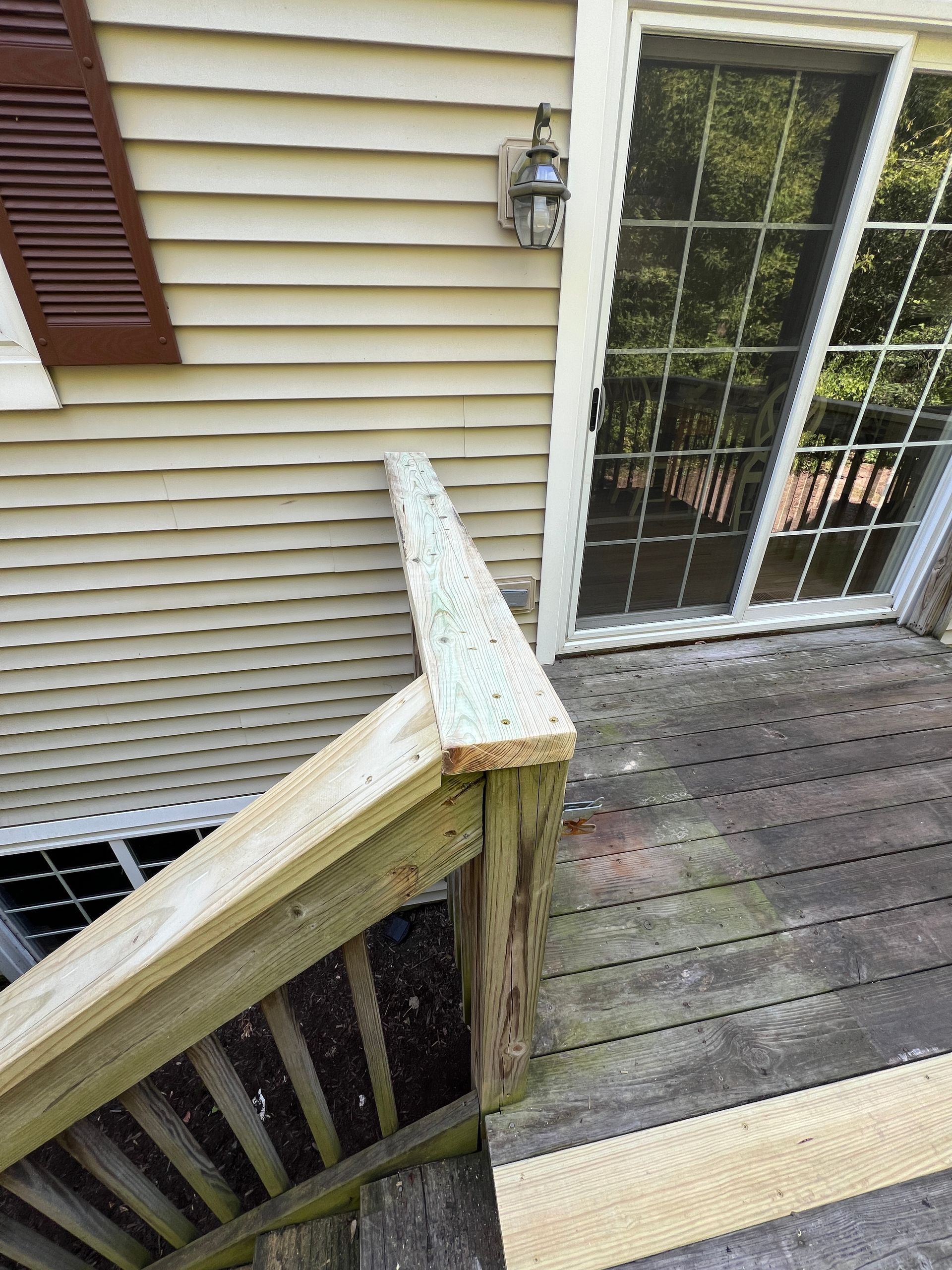 A wooden handrail on a deck staircase leads to a sliding glass door on the side of a house with light-colored siding.