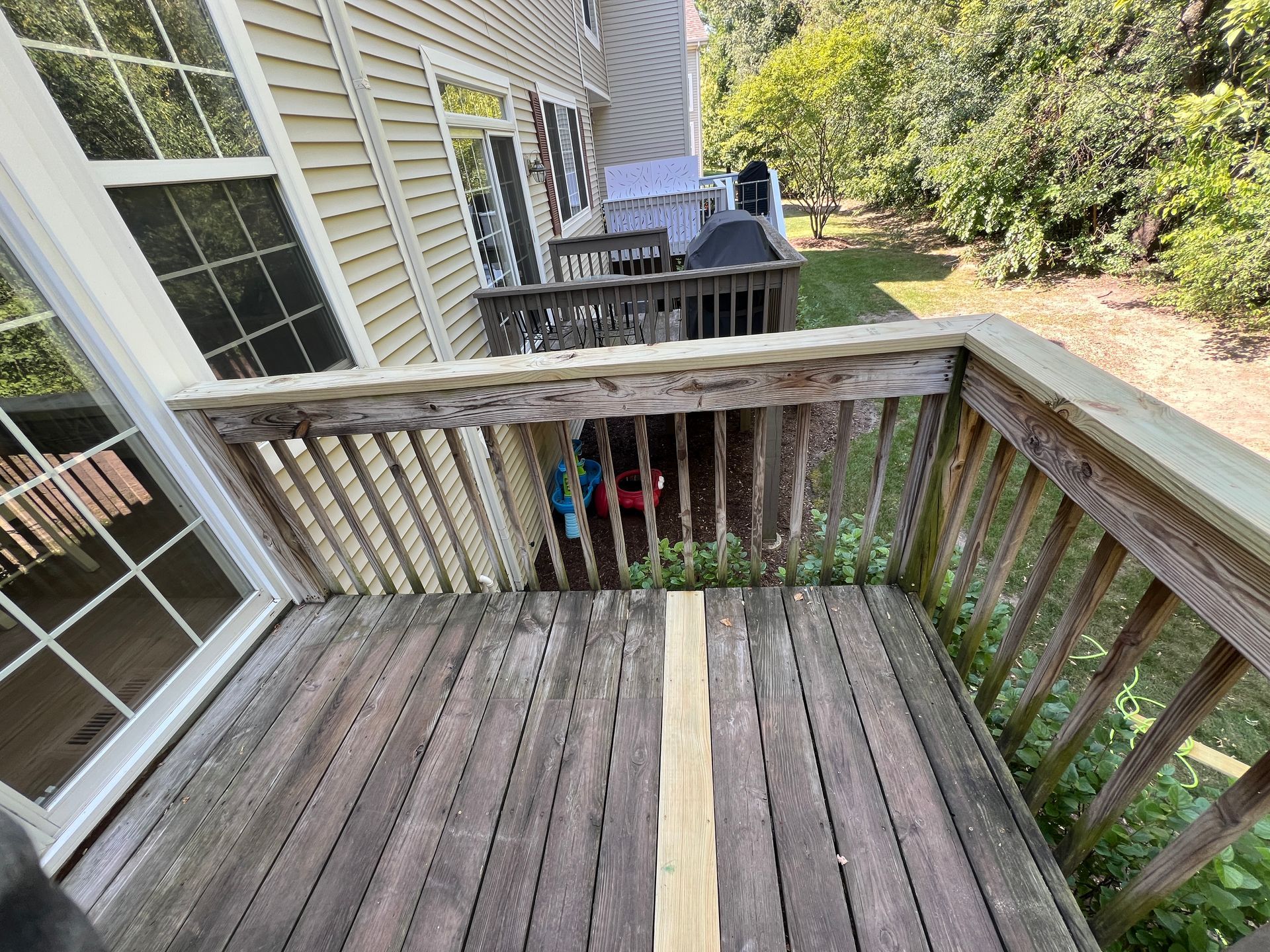 A wooden deck with a newly replaced center board looking out onto a backyard with trees and a second-level balcony.