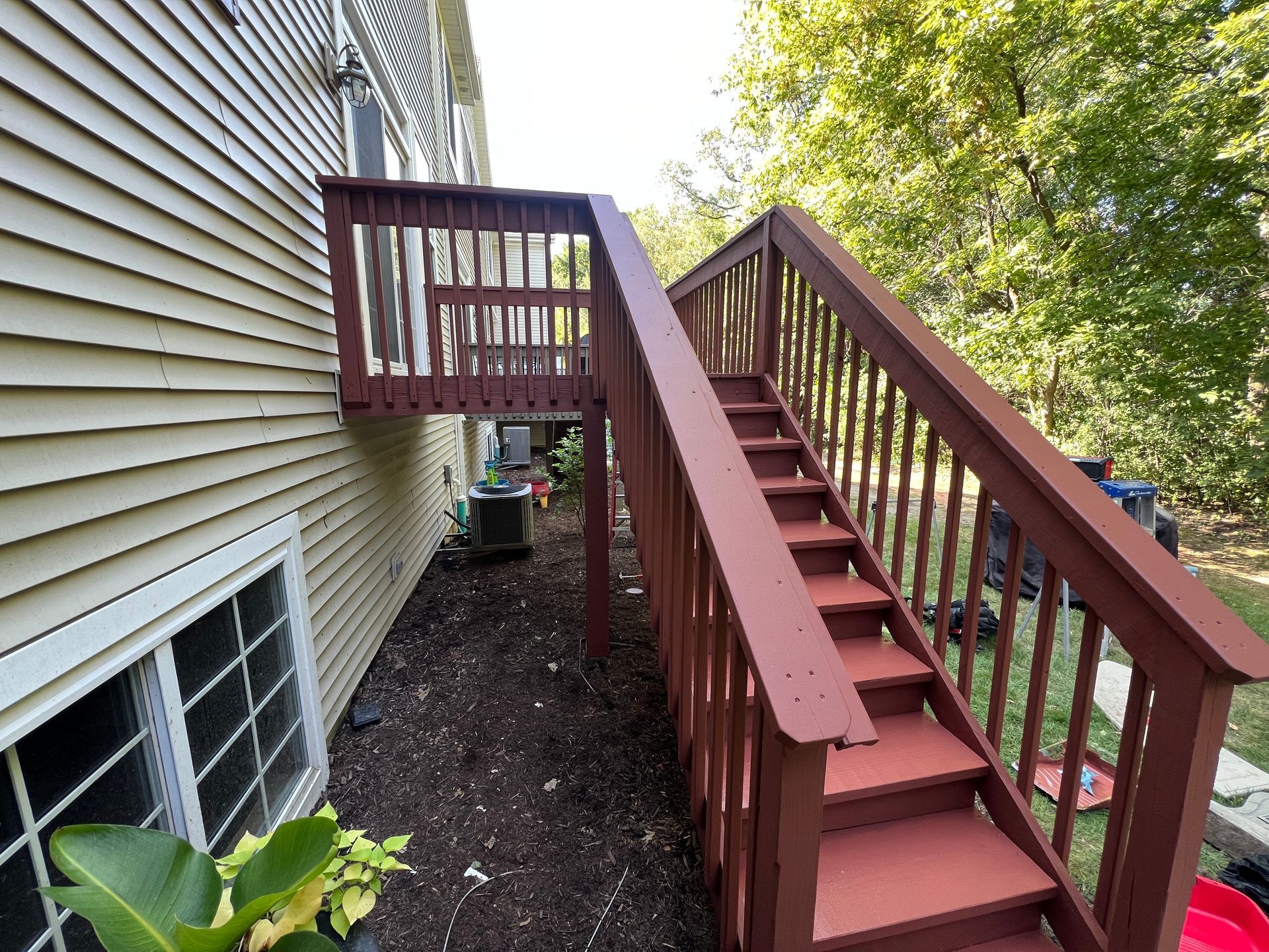 A side view of a house with beige vinyl siding and a dark red wooden deck featuring stairs leading down to a garden bed.