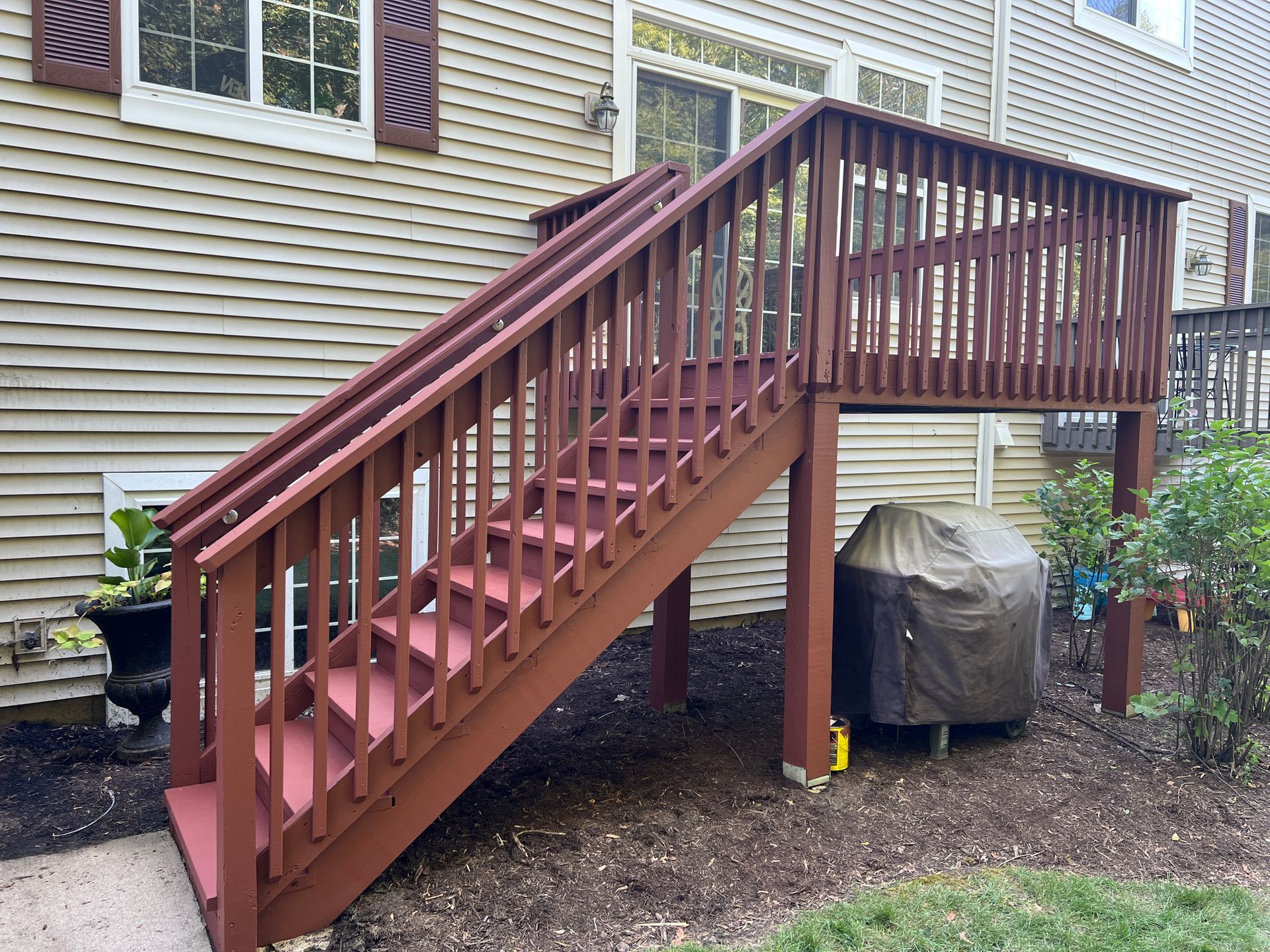 Reddish-brown wooden stairs leading up to an elevated backyard deck against a house with light tan siding.