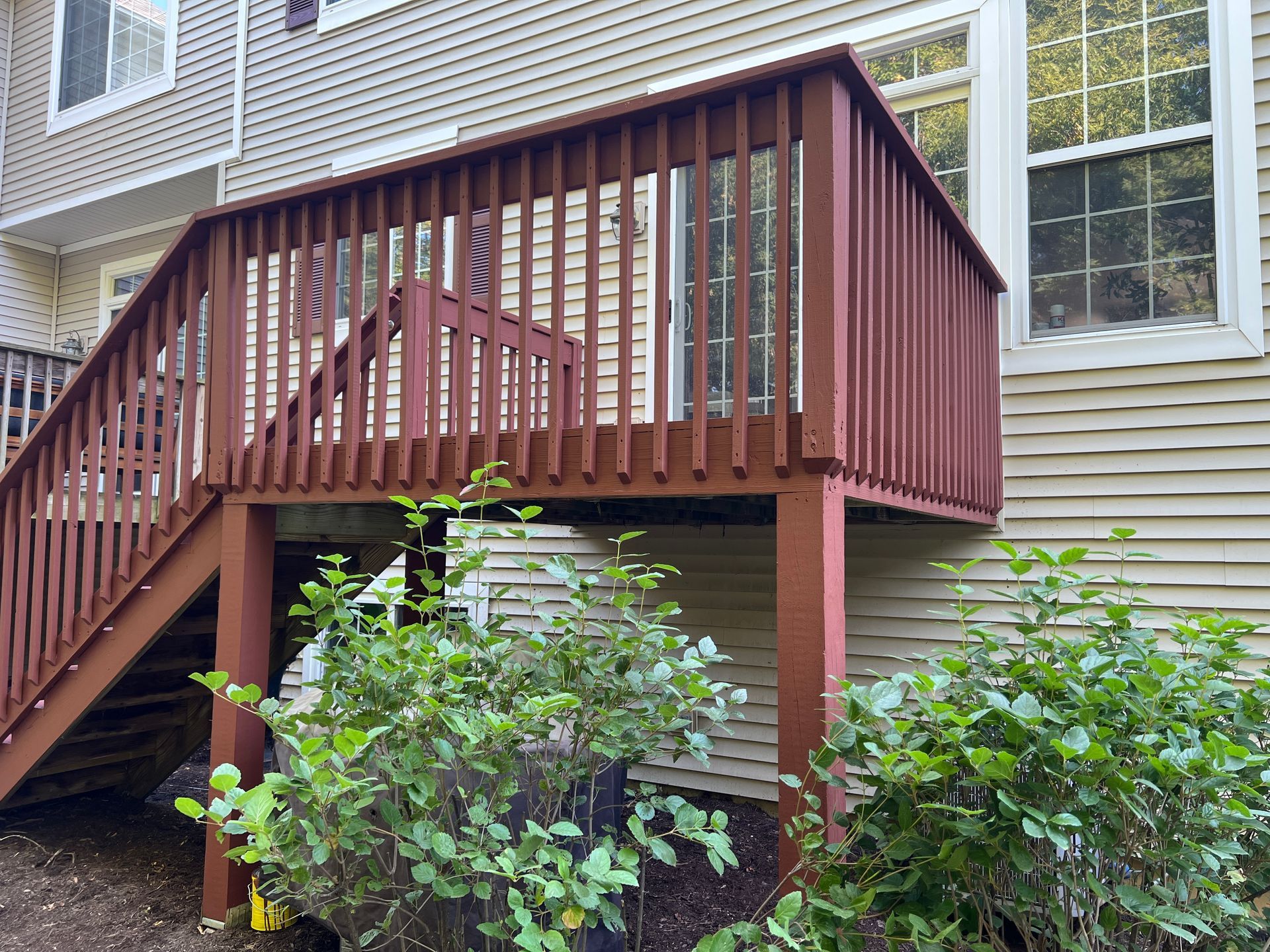 A brown wooden deck with stairs attached to the back of a house with light-colored siding, surrounded by green shrubs.