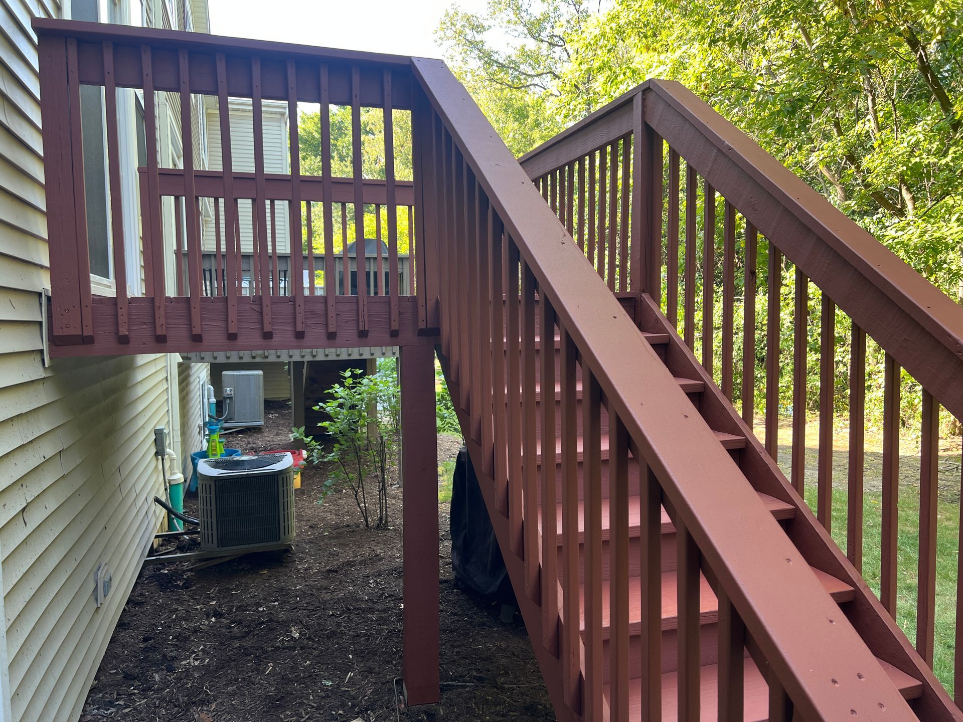 A brown wooden deck with stairs descending to a mulched ground area next to a beige-sided house.