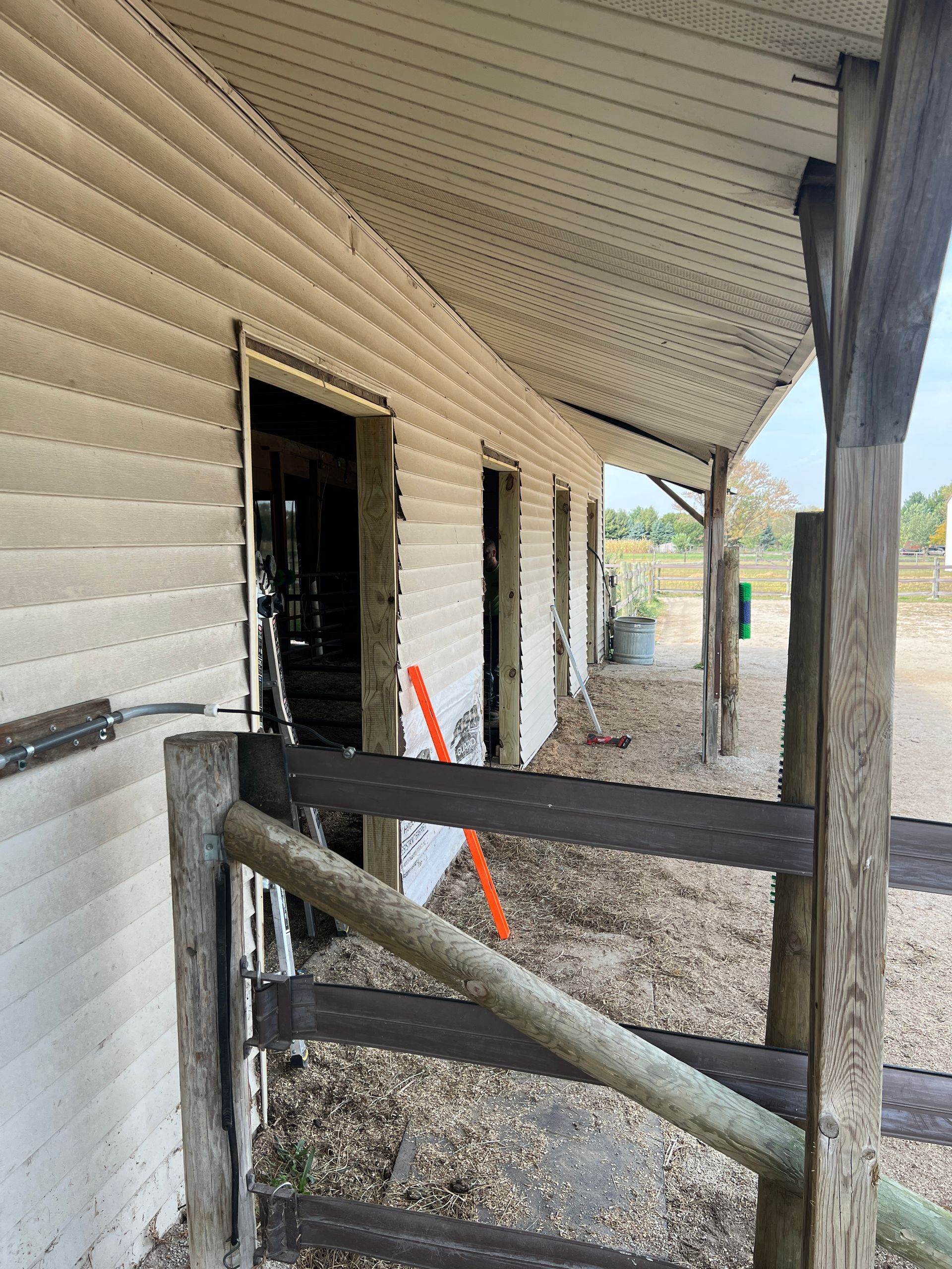 A row of horse stalls with missing doors along a light-colored siding building, partially obscured by a wooden fence.