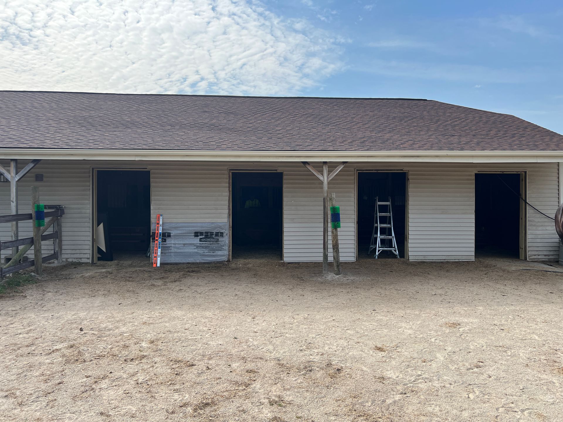 A white horse barn with four open stalls, a brown metal roof, and a gravel yard under a bright, partly cloudy sky.